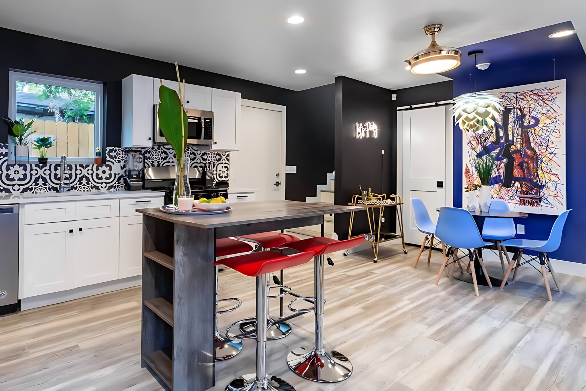 Open-concept kitchen with white cabinets, stainless steel appliances, and a patterned backsplash, featuring a kitchen island with red bar stools, flowing into a dining area with a black table and light blue chairs.