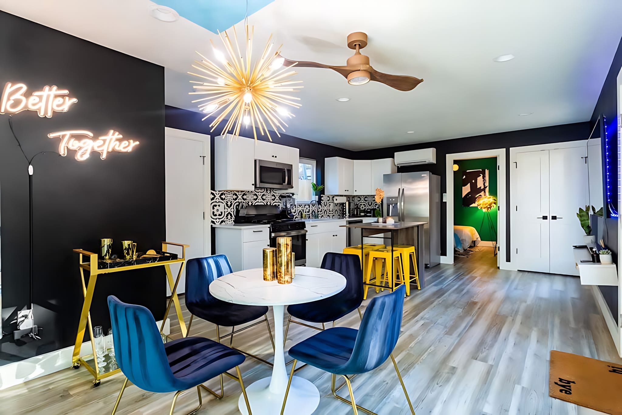 Modern open-concept dining area with a white round table and blue velvet chairs, set against a black accent wall with a "Better Together" neon sign, adjacent to a white kitchen with patterned backsplash.