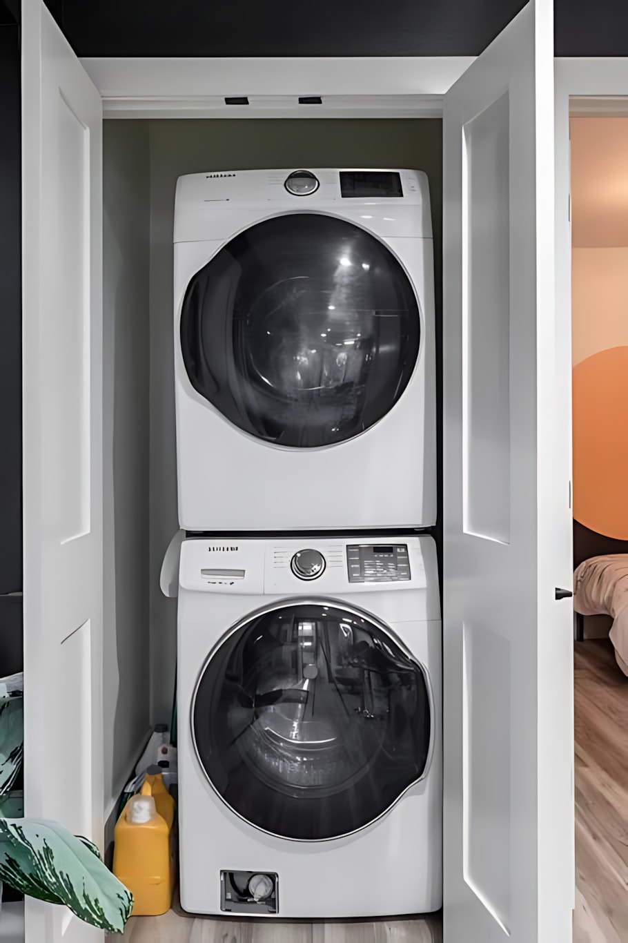 A compact laundry closet featuring a white stacked front-loading washer and dryer unit, with laundry detergent bottles visible on the floor. The appliances are neatly tucked behind white bi-fold doors.