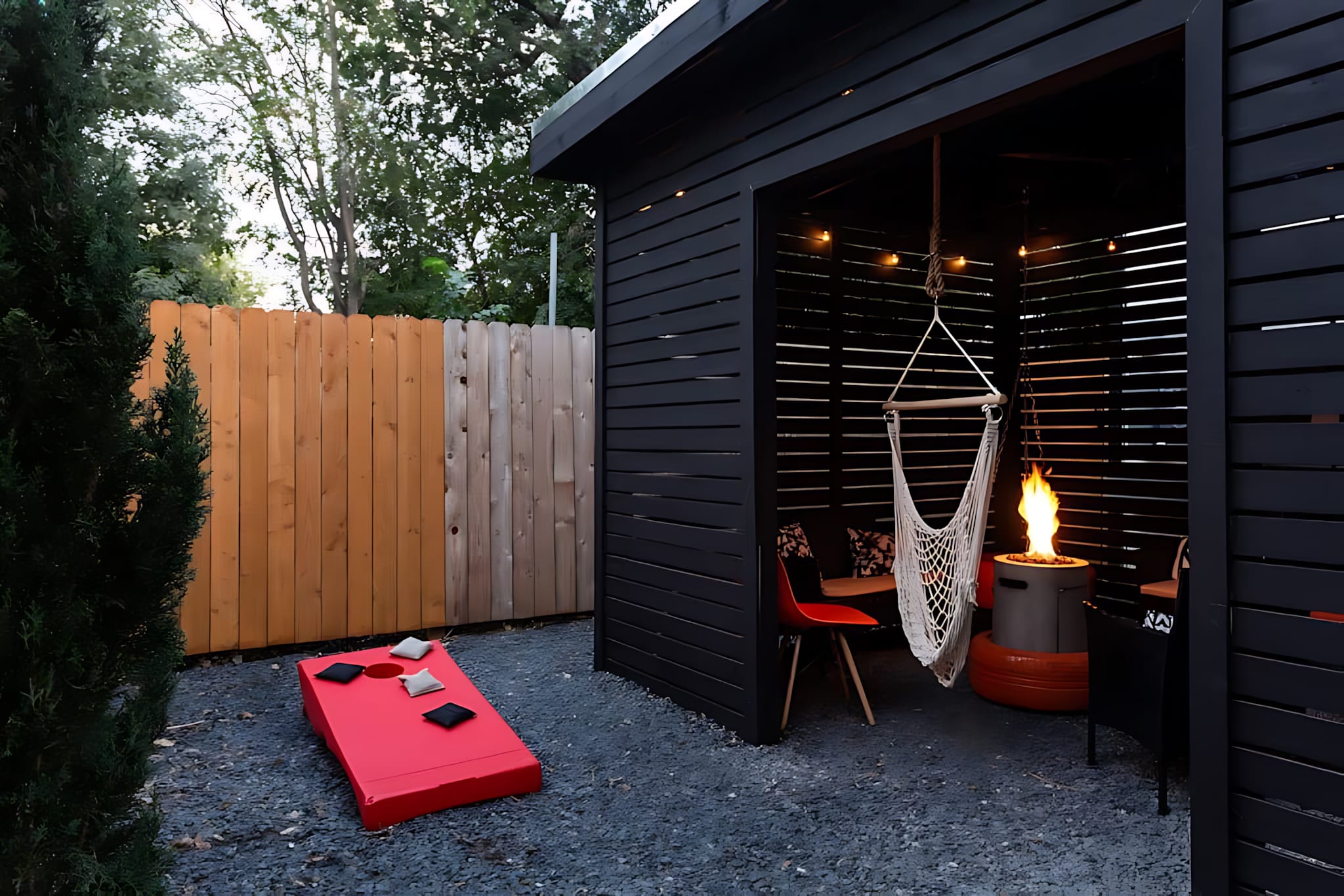 A gravel backyard area featuring a black slatted shed with a hanging hammock chair and a fire pit, alongside a red cornhole game set and a wooden fence. String lights illuminate the covered outdoor seating area.