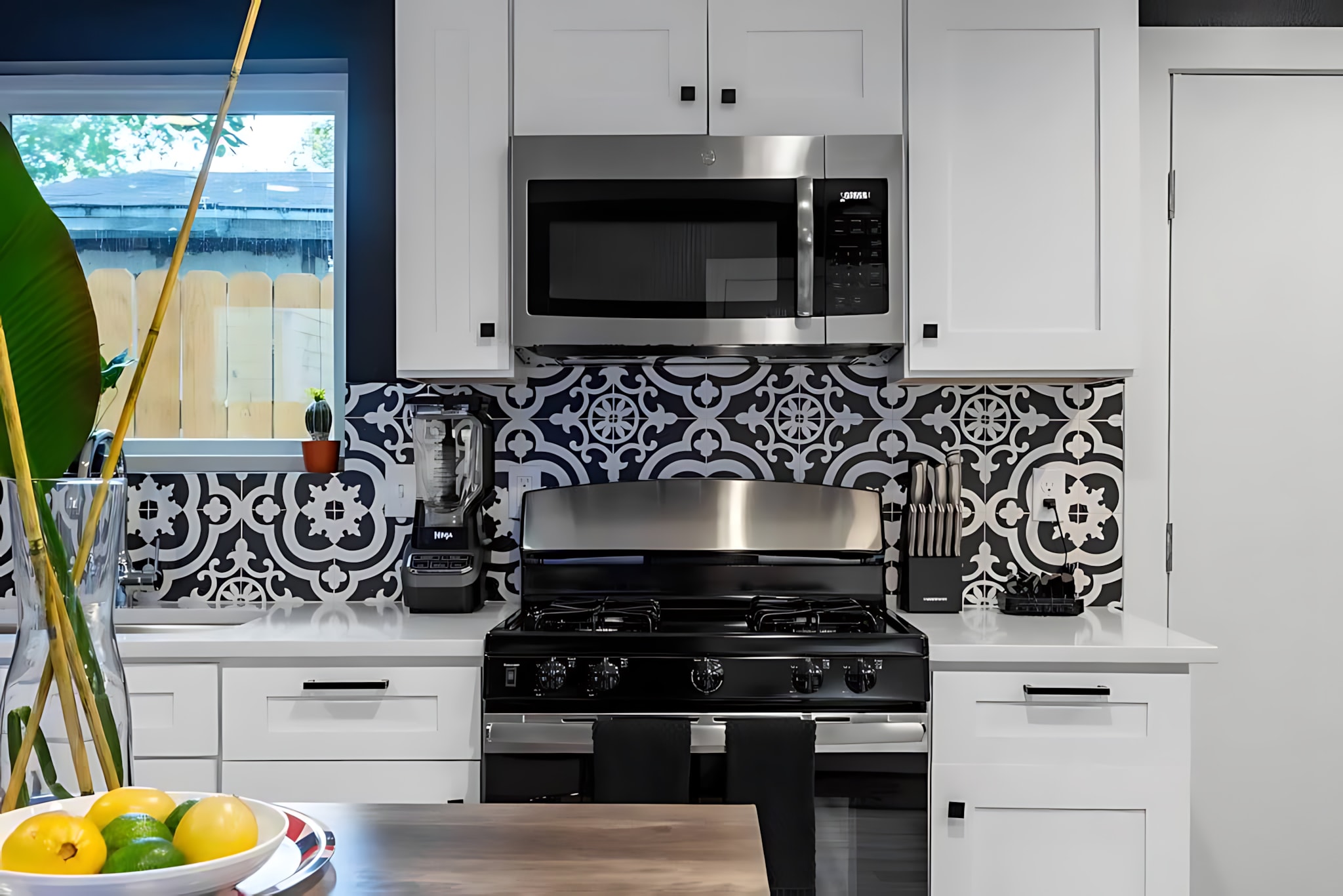Modern kitchen featuring white shaker cabinets, stainless steel appliances, and a striking black and white patterned tile backsplash. A gas range, microwave, and blender are visible on the white countertop.
