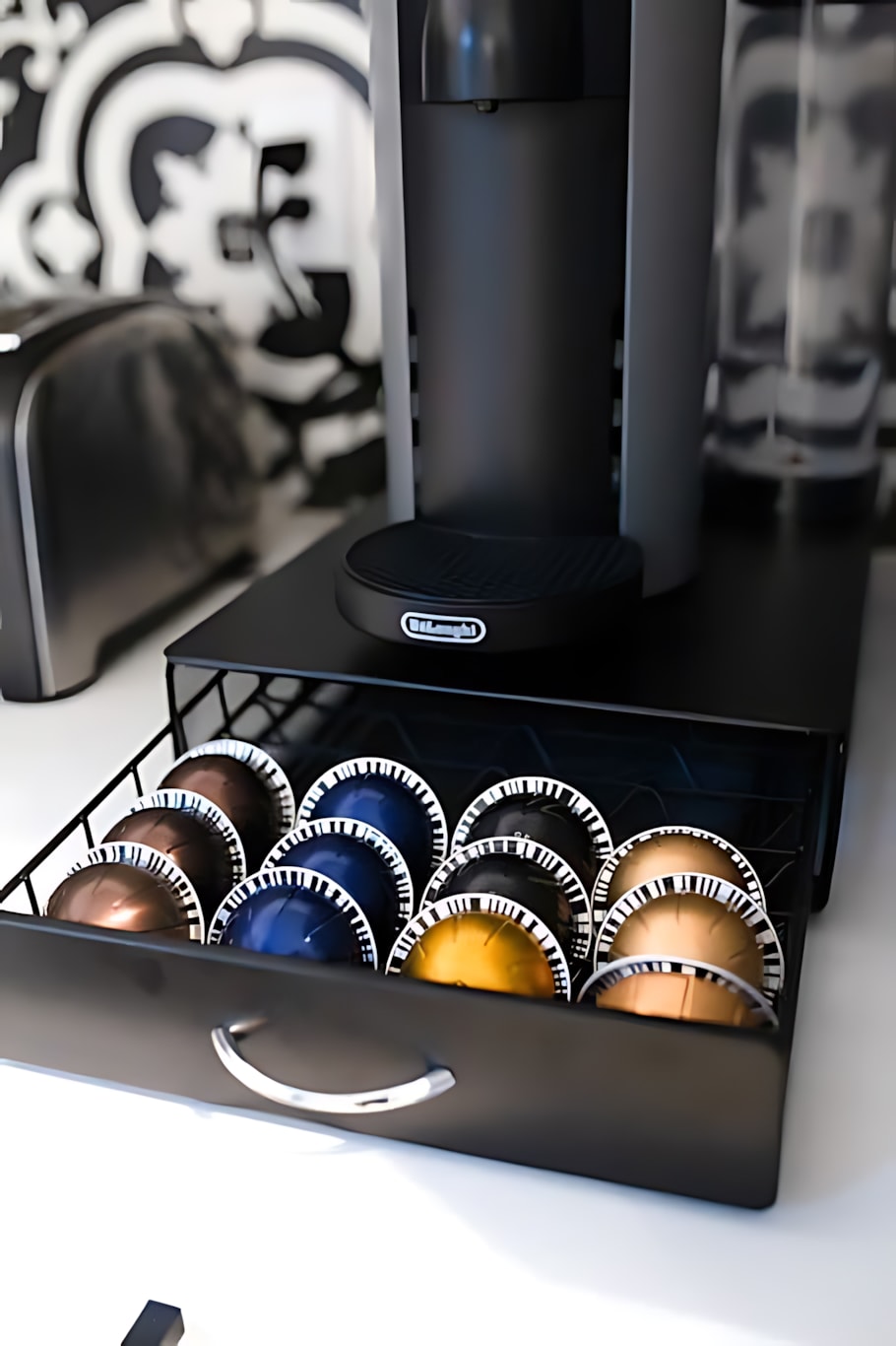 Close-up of a black Nespresso coffee machine sitting on a black stand with a pull-out drawer filled with various colored coffee pods. The patterned tile backsplash is visible in the blurred background.