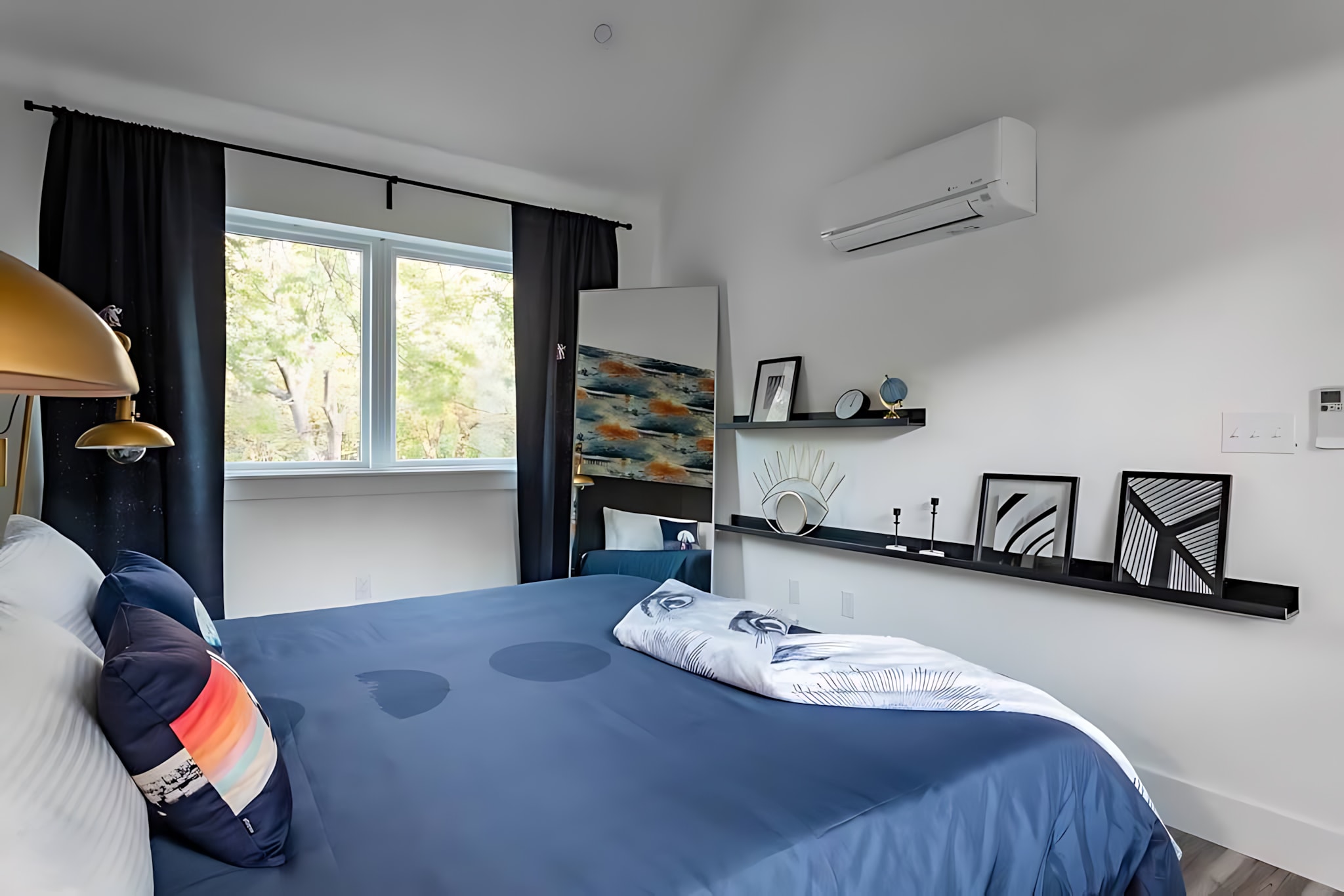 A contemporary bedroom with a blue-covered bed, black curtains framing a large window, and a white wall adorned with black floating shelves displaying decorative items.