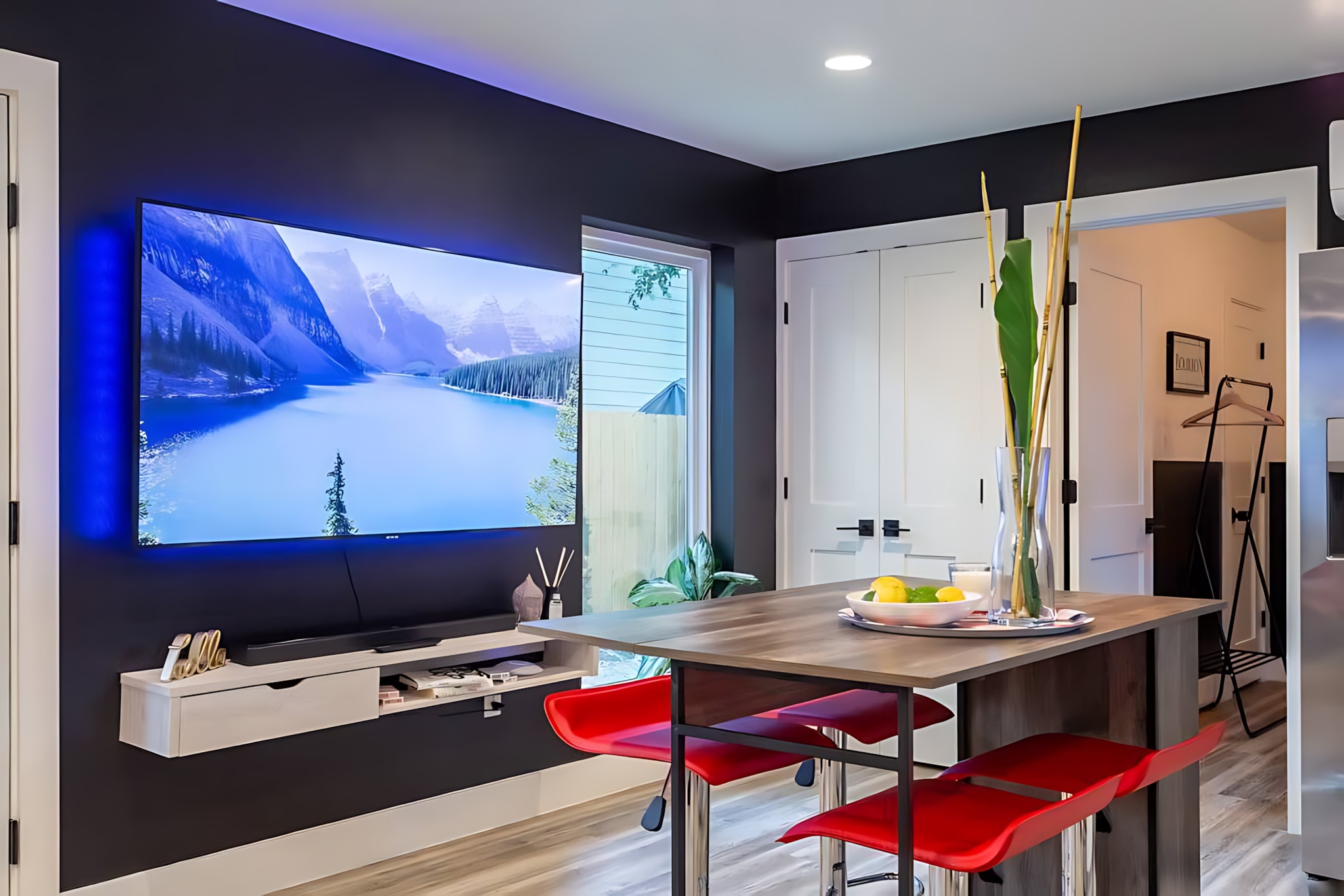 A modern living area featuring a large wall-mounted TV with blue backlighting against a dark accent wall. A wooden island with red bar stools serves as a dining or casual seating area, with a window offering an outdoor view.