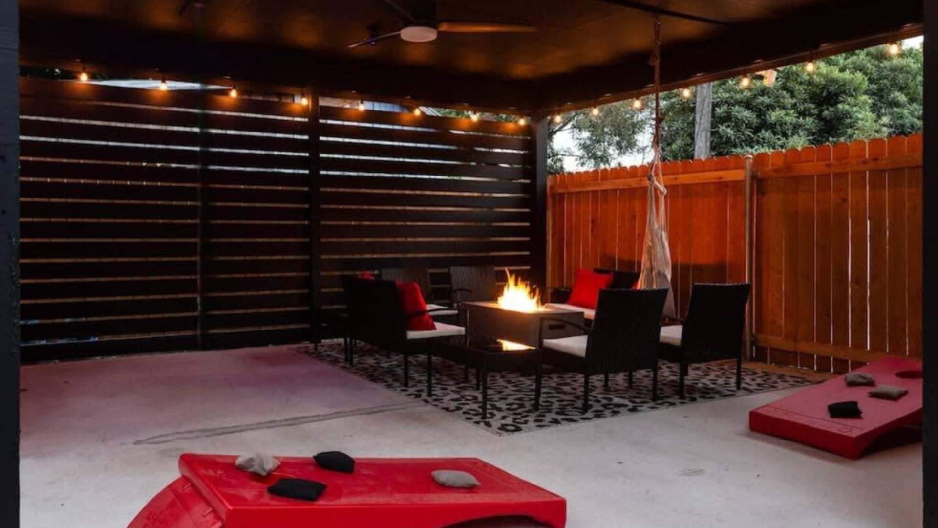 Spacious covered patio featuring a central fire pit, black outdoor seating with white cushions, a hanging hammock chair, and string lights, with cornhole boards visible on the concrete ground.