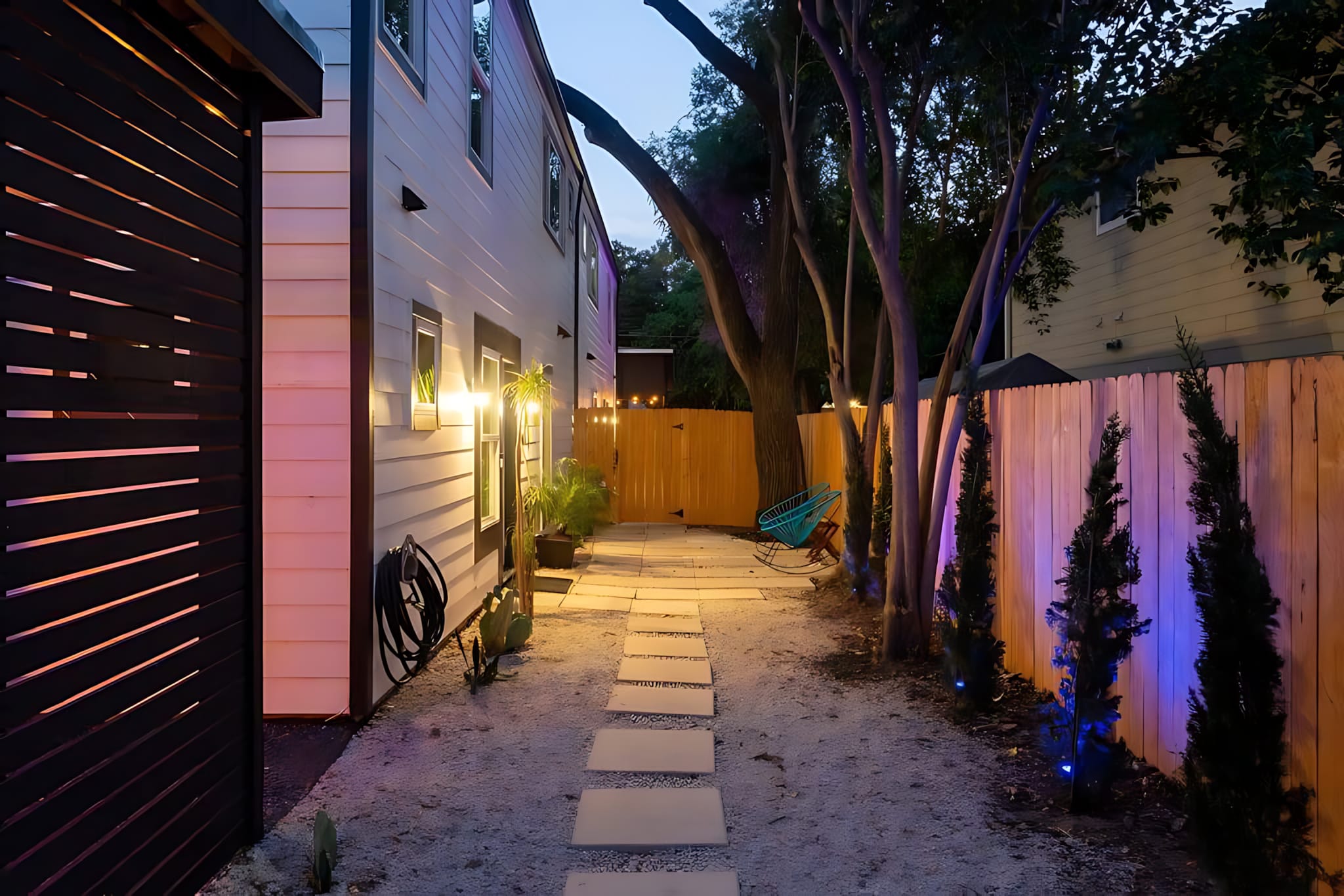 A gravel pathway with square stepping stones leads through a narrow backyard, flanked by a white-sided house with wall lights on the left and a wooden fence with trees and blue-lit shrubs on the right.