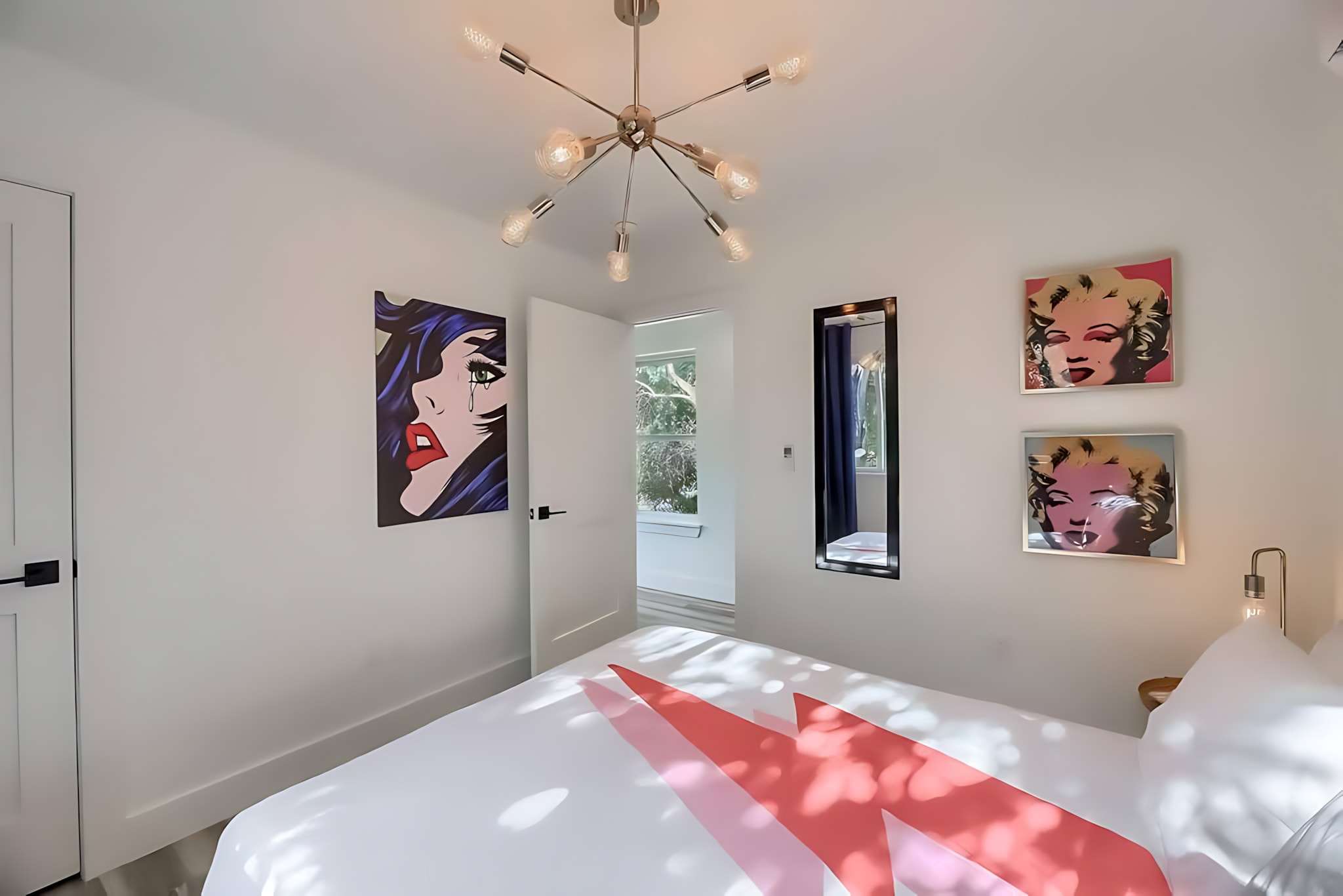 A bright bedroom with a queen bed, pop art on the walls including a crying woman and Marilyn Monroe prints, and a modern sputnik-style ceiling light fixture. An open door reveals an outdoor view.