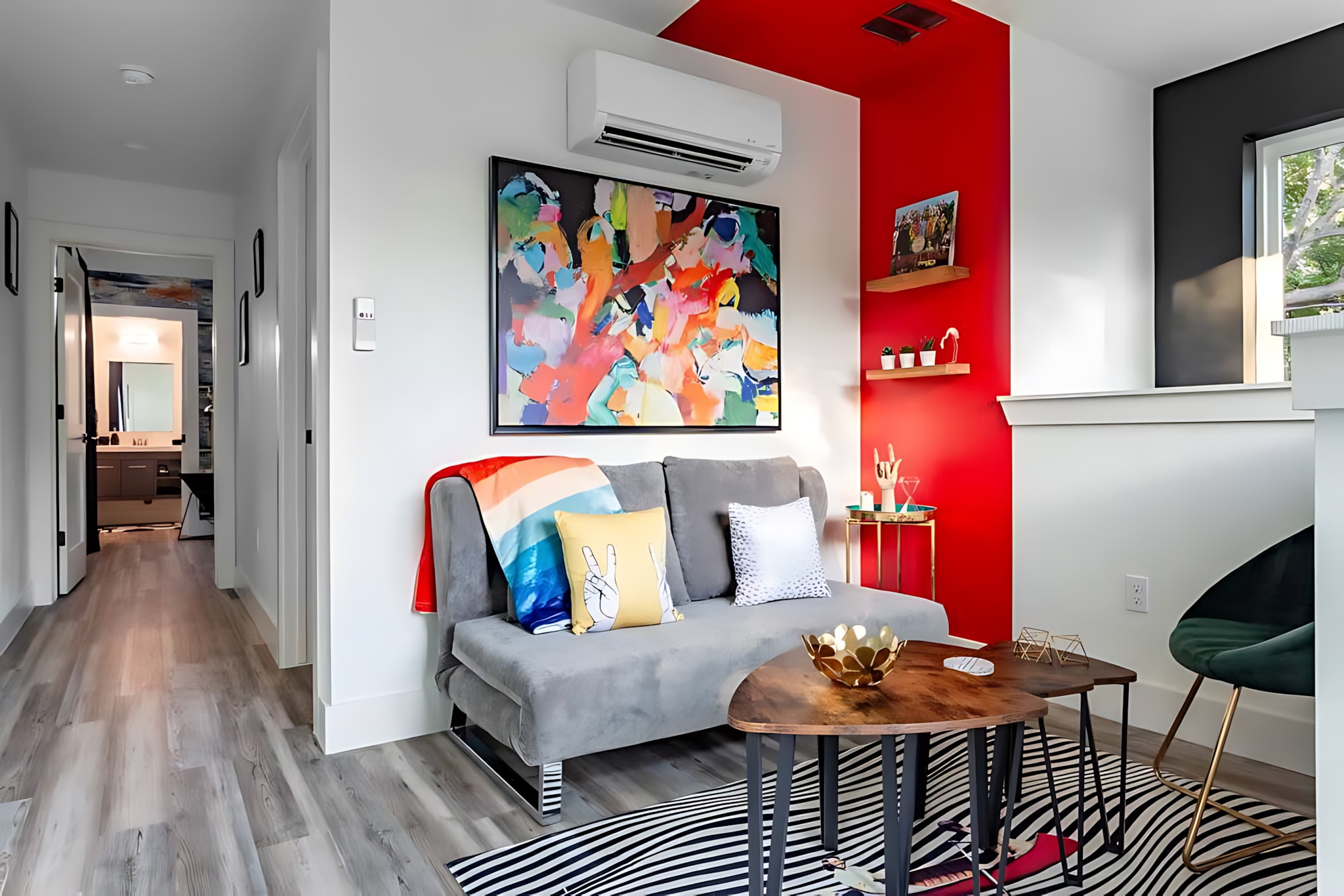 A vibrant living area with a grey chaise lounge, colorful throw pillows, and a large abstract art piece above. A red accent wall adds a pop of color, and a multi-level coffee table sits on a black and white striped rug.