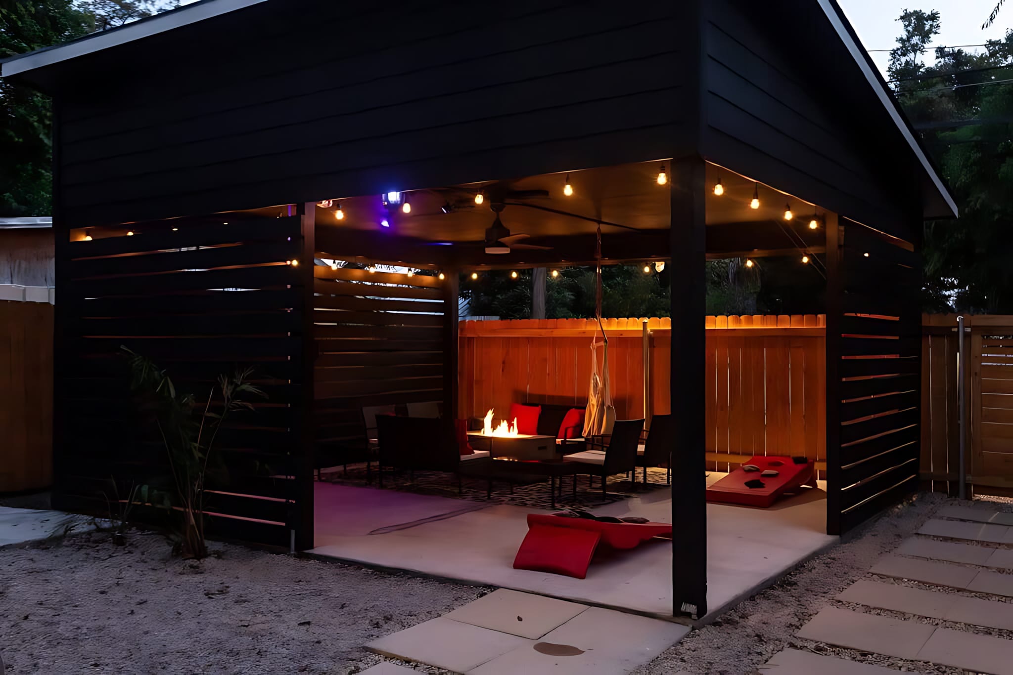 A modern black slatted patio structure with string lights illuminates a central fire pit, surrounded by dark outdoor seating and red cushions, creating a cozy evening ambiance.
