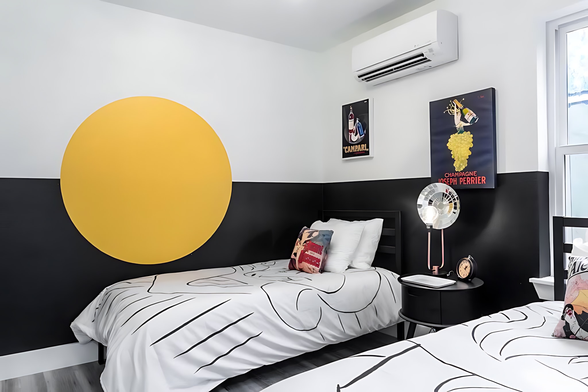 A modern bedroom with a black and white color scheme, featuring two twin beds with graphic bedding, and a large yellow circular art piece on the wall. Vintage posters adorn the space, and an AC unit is mounted high on the wall.