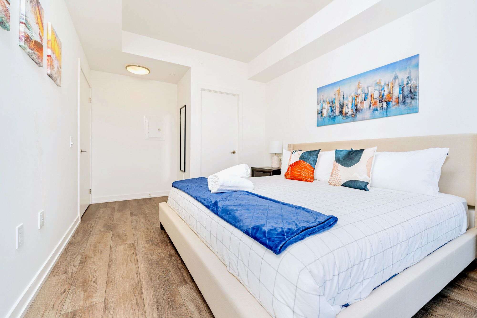 Spacious bedroom with light wood-look flooring, a large bed with a beige headboard, white and blue bedding, and modern artwork above the bed.