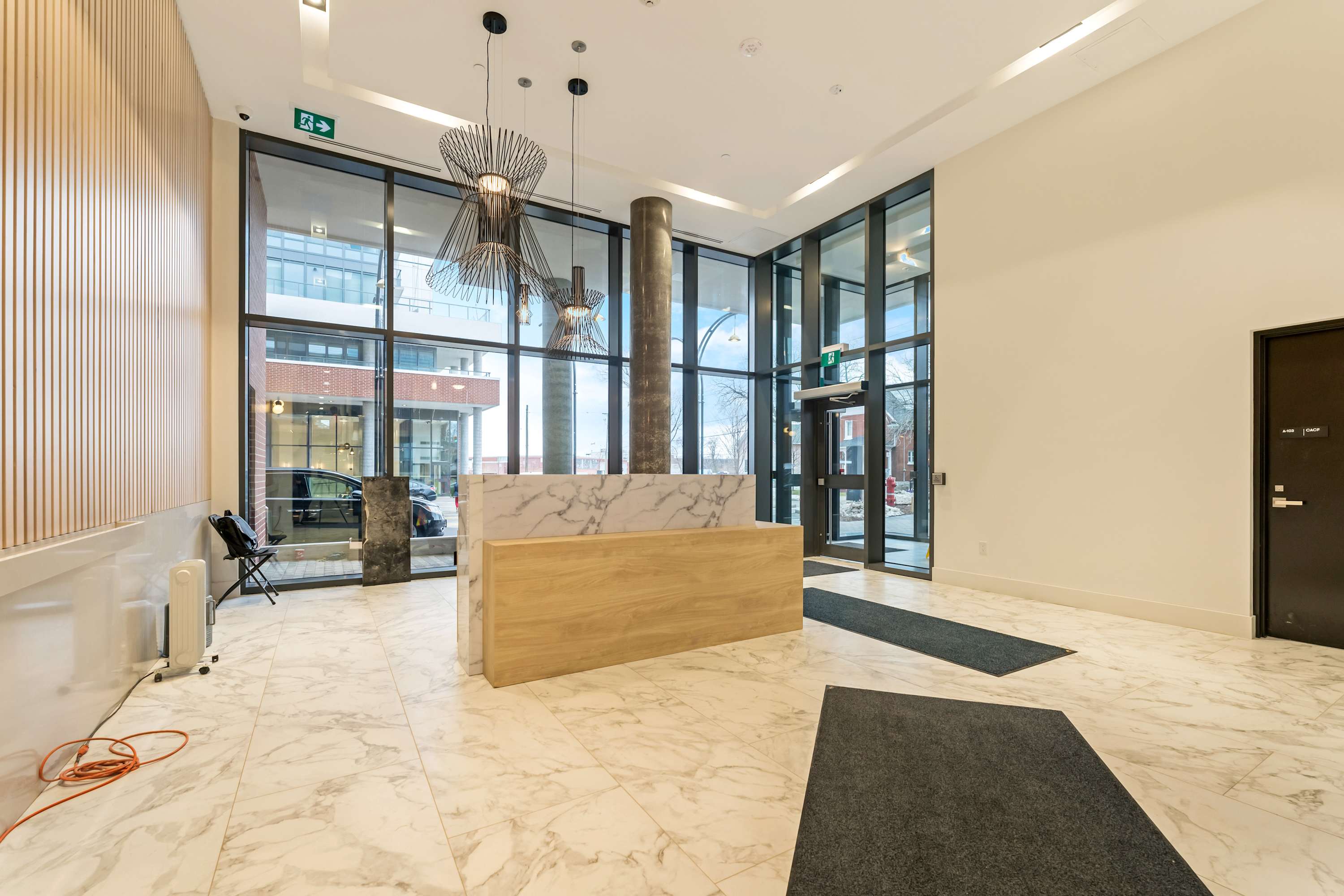 A bright and modern building lobby features a reception desk with a white marble top and a light wood base, set against a backdrop of large floor-to-ceiling windows. The floor is covered in white marble tiles with grey veining, and contemporary pendant lights hang from the ceiling.