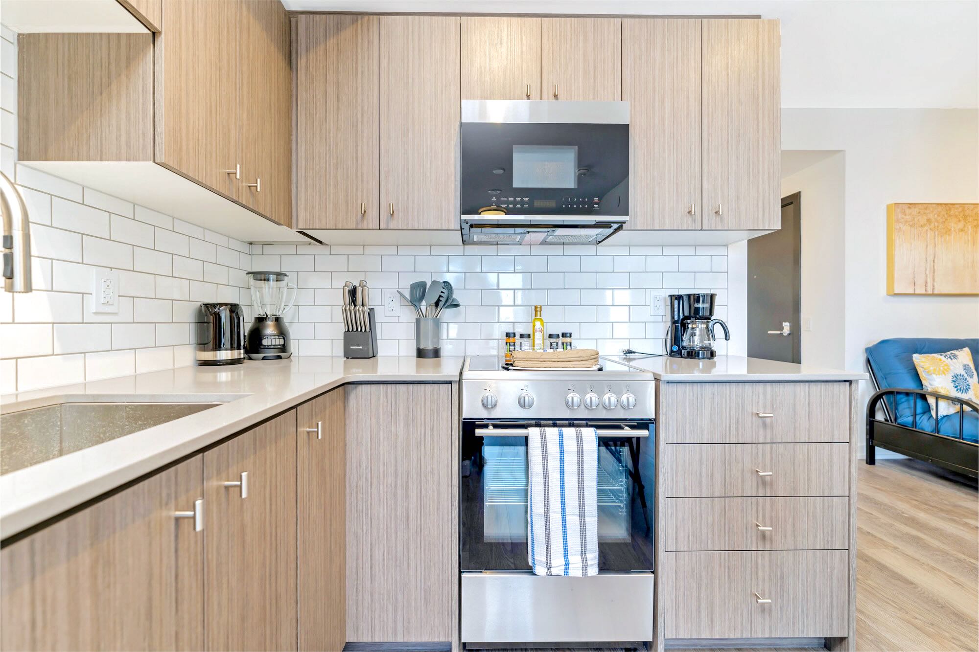 Corner view of a modern kitchen featuring light wood cabinetry, white subway tile backsplash, stainless steel oven/range, and a microwave above, with various small appliances on the light countertop.