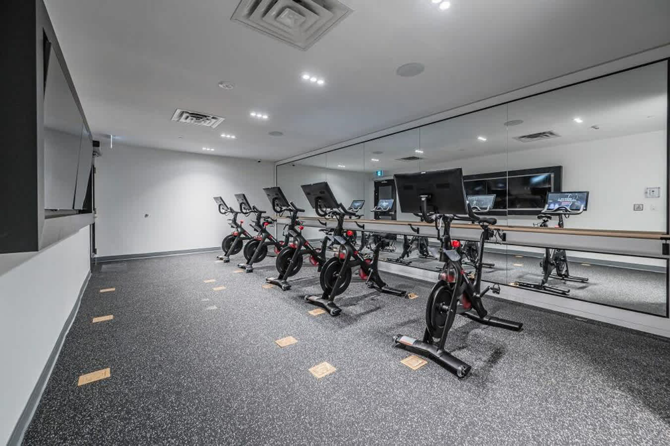 A modern fitness room features a row of six black spin bikes facing a large mirrored wall, with individual screens on each bike. The room has a dark speckled rubber floor with light square accents and recessed lighting in the white ceiling.