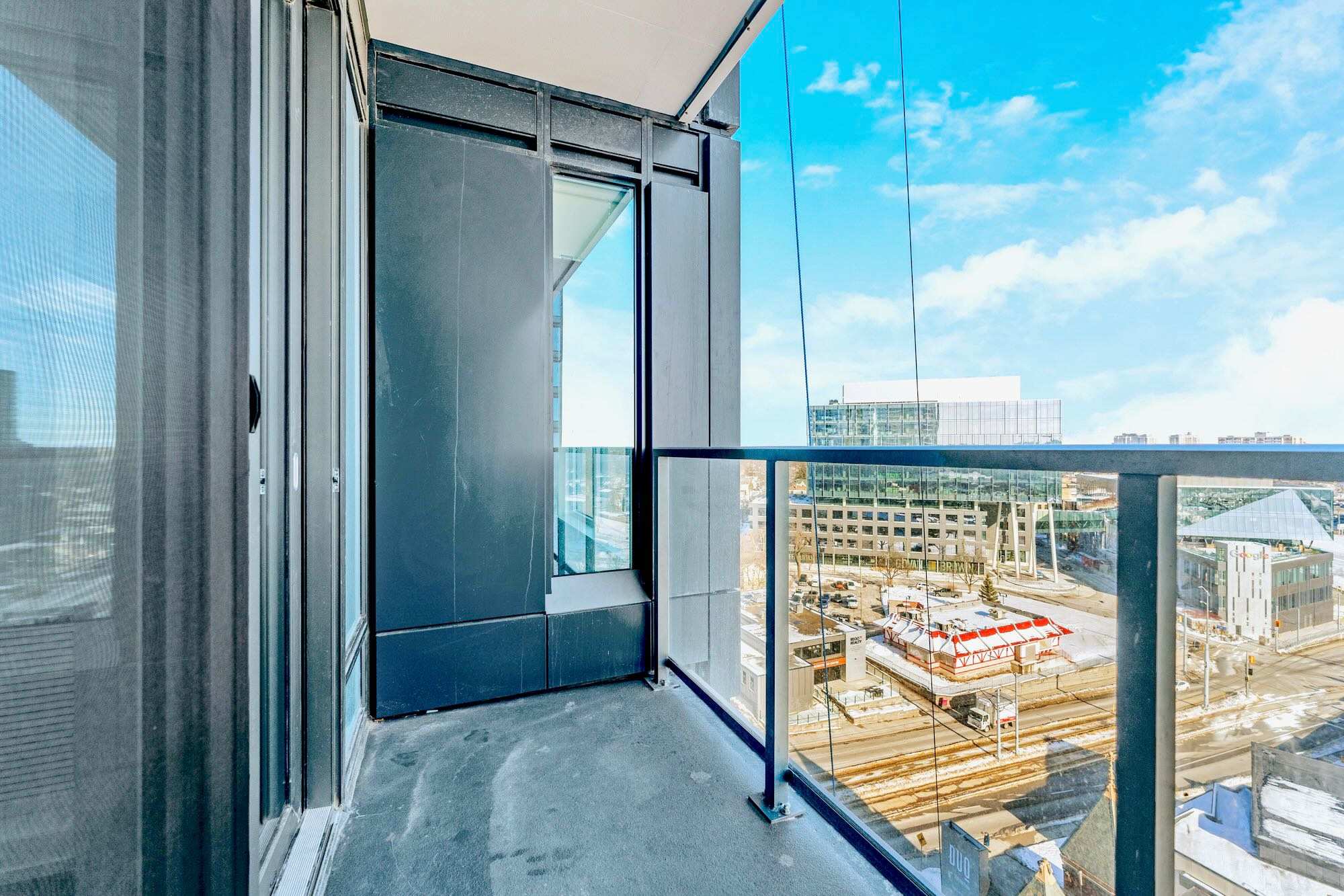 Corner balcony with glass railings providing a wide view of the city below, including commercial buildings and a snowy ground. The modern black exterior of the building is visible on the left.