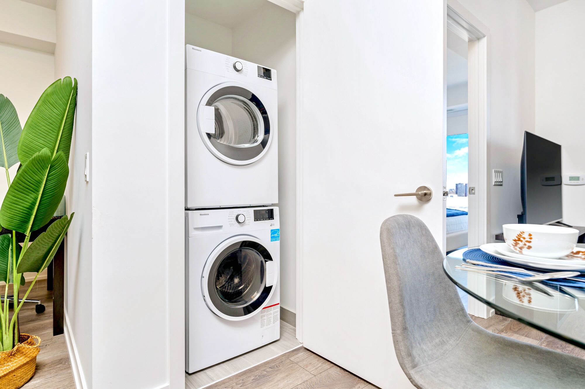 Stacked white washer and dryer units located in a closet, adjacent to a dining area, with light wood-look flooring.