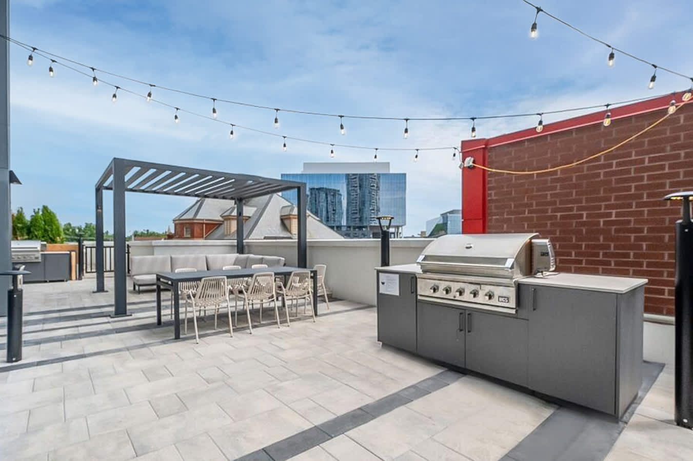 A spacious rooftop patio features a modern grey pergola over a dining table and sofa seating, alongside a large outdoor kitchen with a built-in grill. String lights hang overhead, and the paved surface has dark grey accents, with city buildings visible in the background.