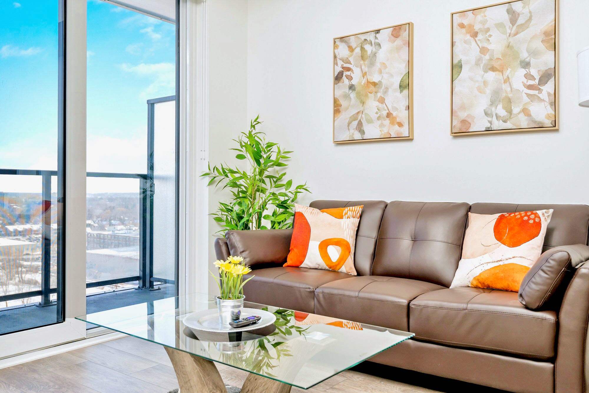 Bright living room featuring a brown leather sofa with orange accent pillows, a glass coffee table, and a green plant. Sliding glass doors lead to a balcony with city views.