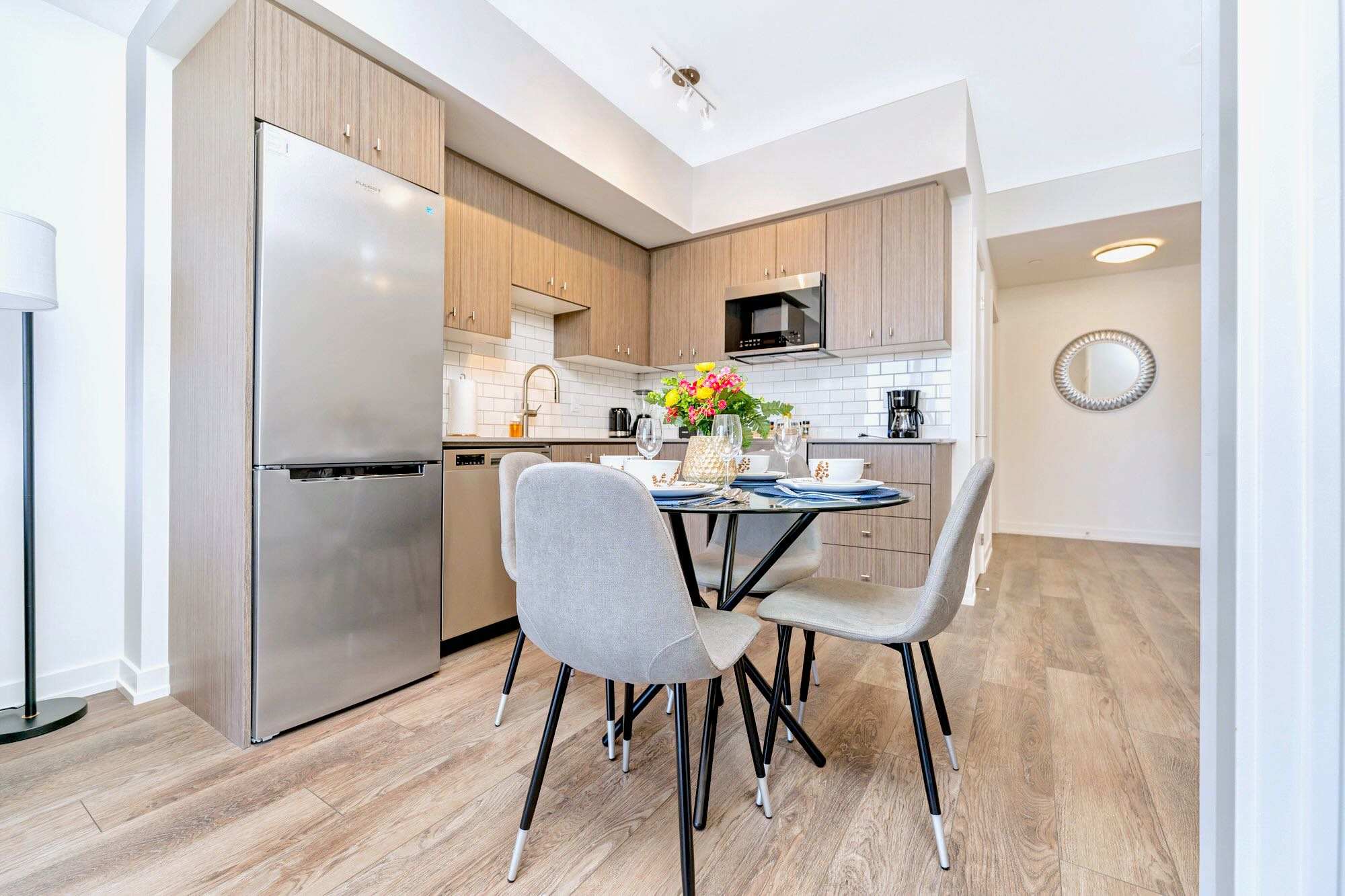 Modern kitchen featuring light wood cabinets, stainless steel refrigerator, microwave, and dishwasher, with a white subway tile backsplash. A round glass dining table with four chairs is positioned in the foreground.