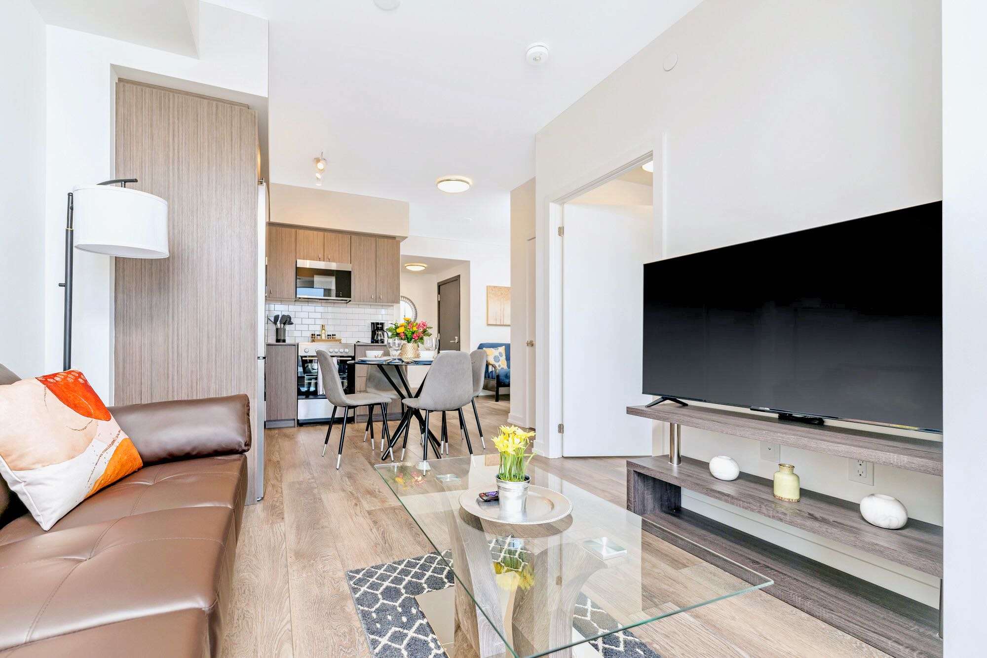 Open concept living area showing the brown leather sofa, glass coffee table, and TV on the left, flowing into the dining area and kitchen on the right. Hardwood floors connect the spaces, and a balcony is visible through the living room windows.