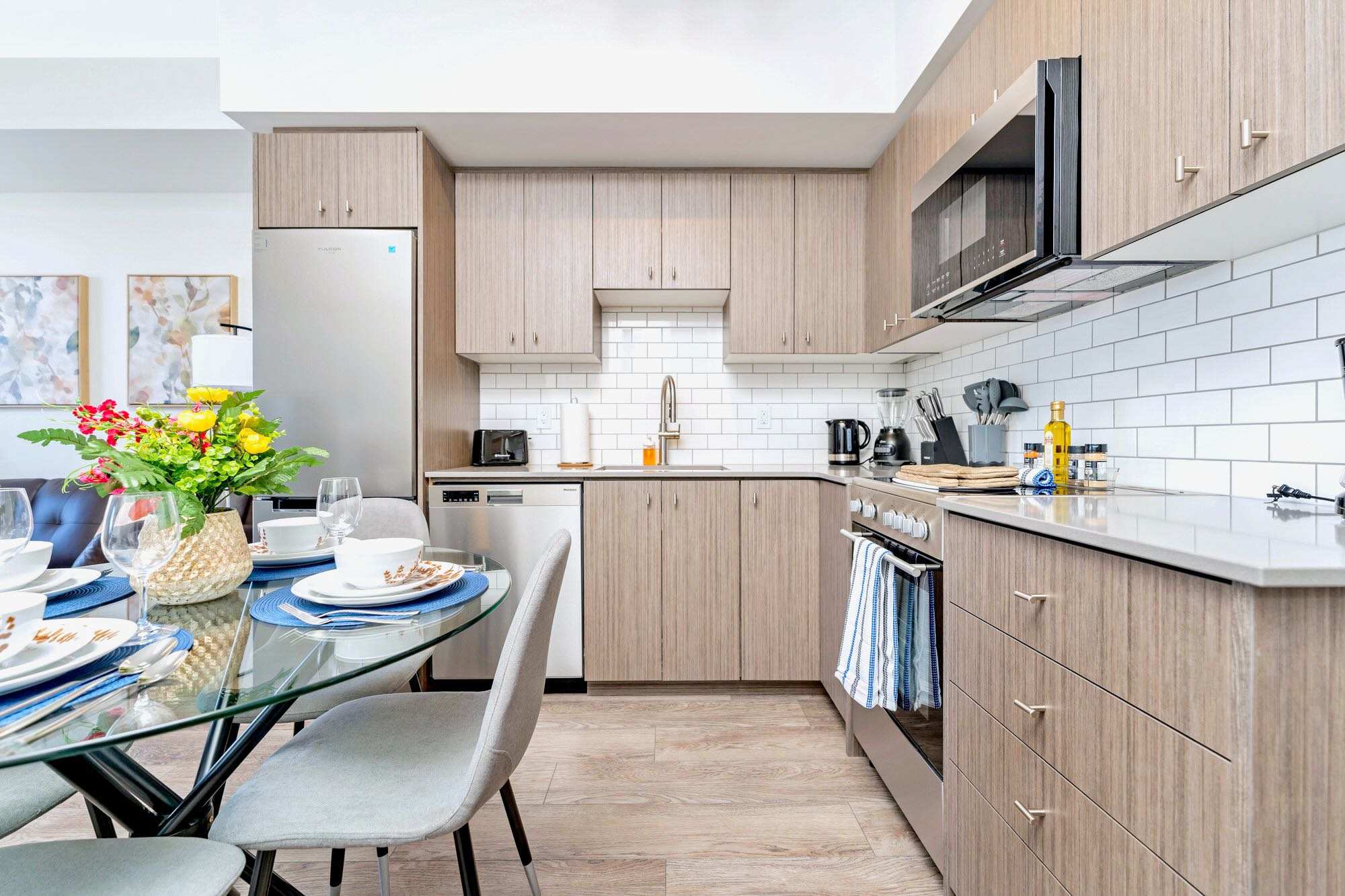 Modern kitchen with light wood cabinetry, white subway tile backsplash, stainless steel appliances, and a glass-top dining table set for four with grey chairs.