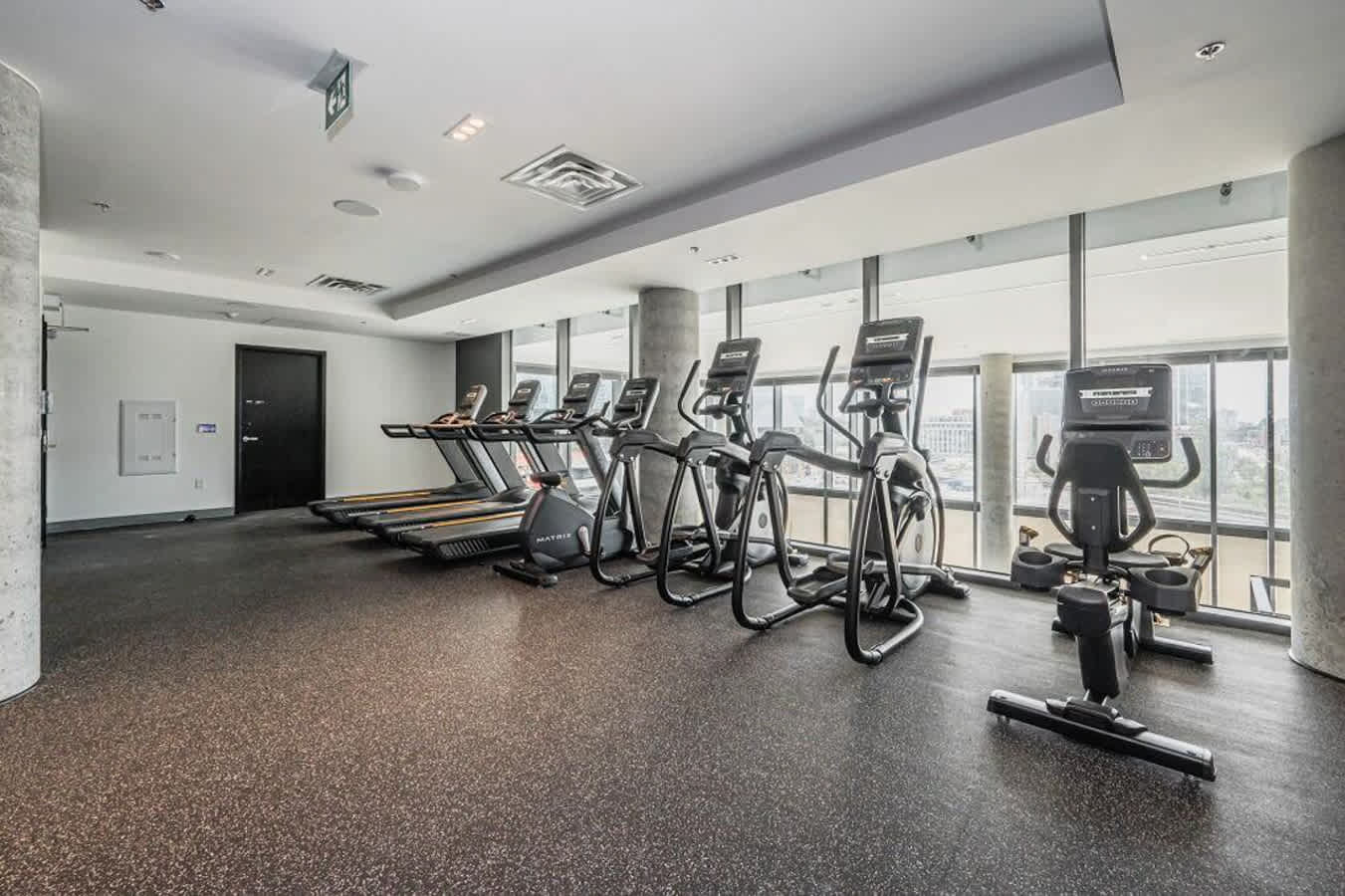 A well-equipped fitness center features several treadmills, elliptical machines, and a stationary bike lined up against large windows offering city views. The room has a dark speckled rubber floor, concrete pillars, and a modern ceiling with recessed lighting.