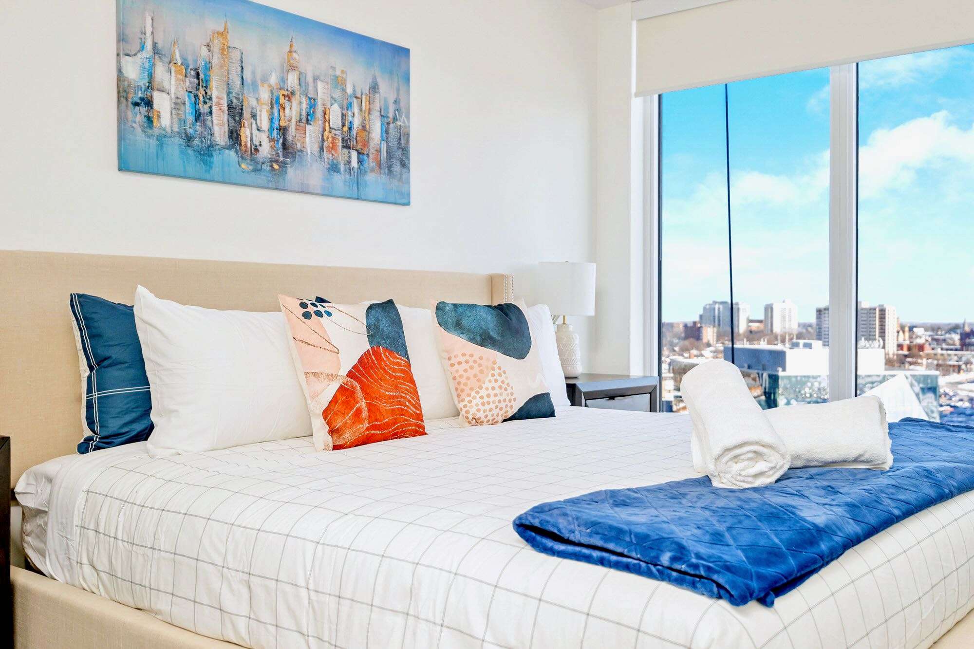 Modern bedroom with a large bed featuring a beige headboard, white and blue bedding, colorful accent pillows, and floor-to-ceiling windows offering city views.