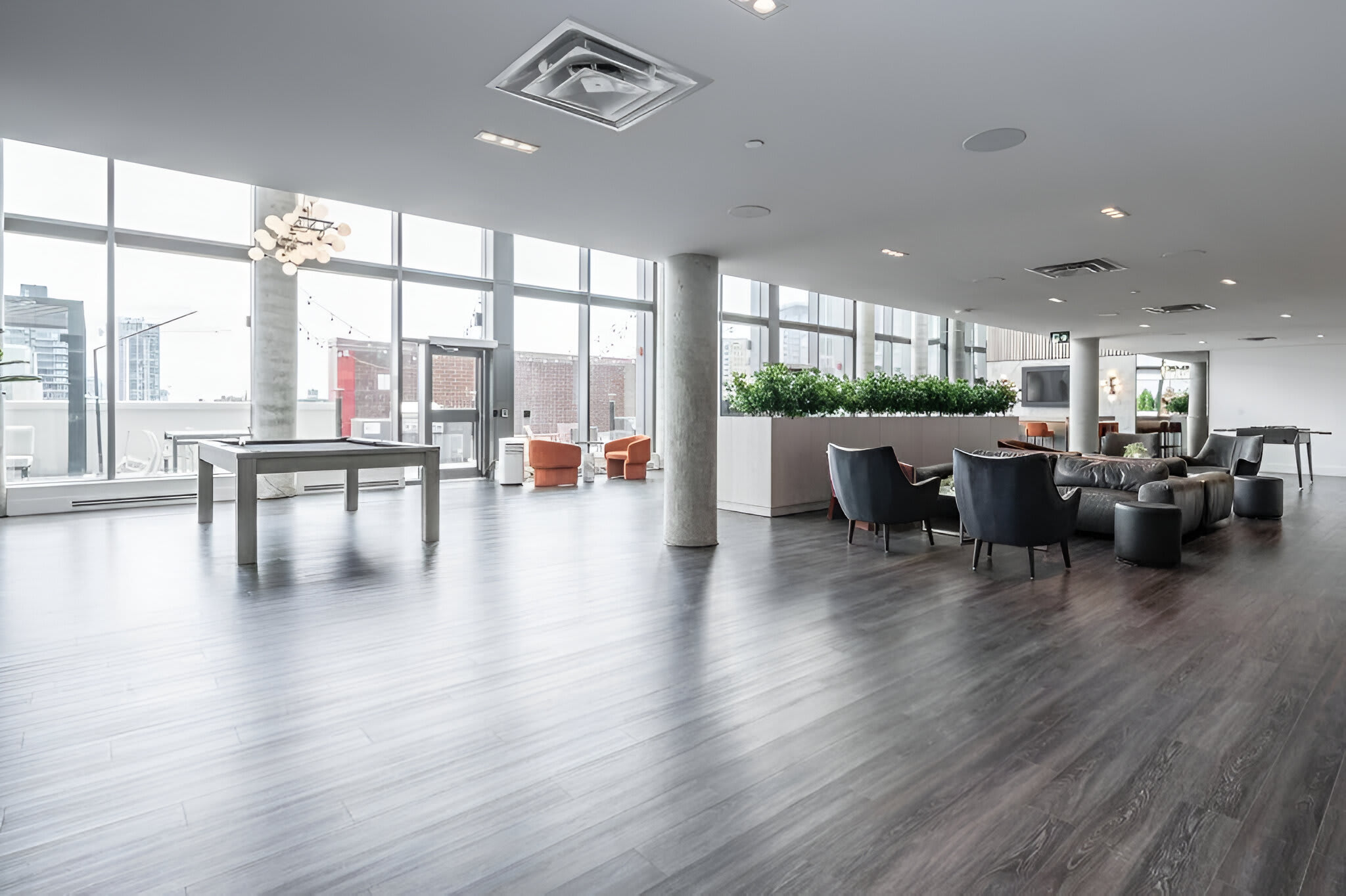 Expansive common area with dark wood-look flooring, a pool table, various lounge seating arrangements, and floor-to-ceiling windows offering city views.