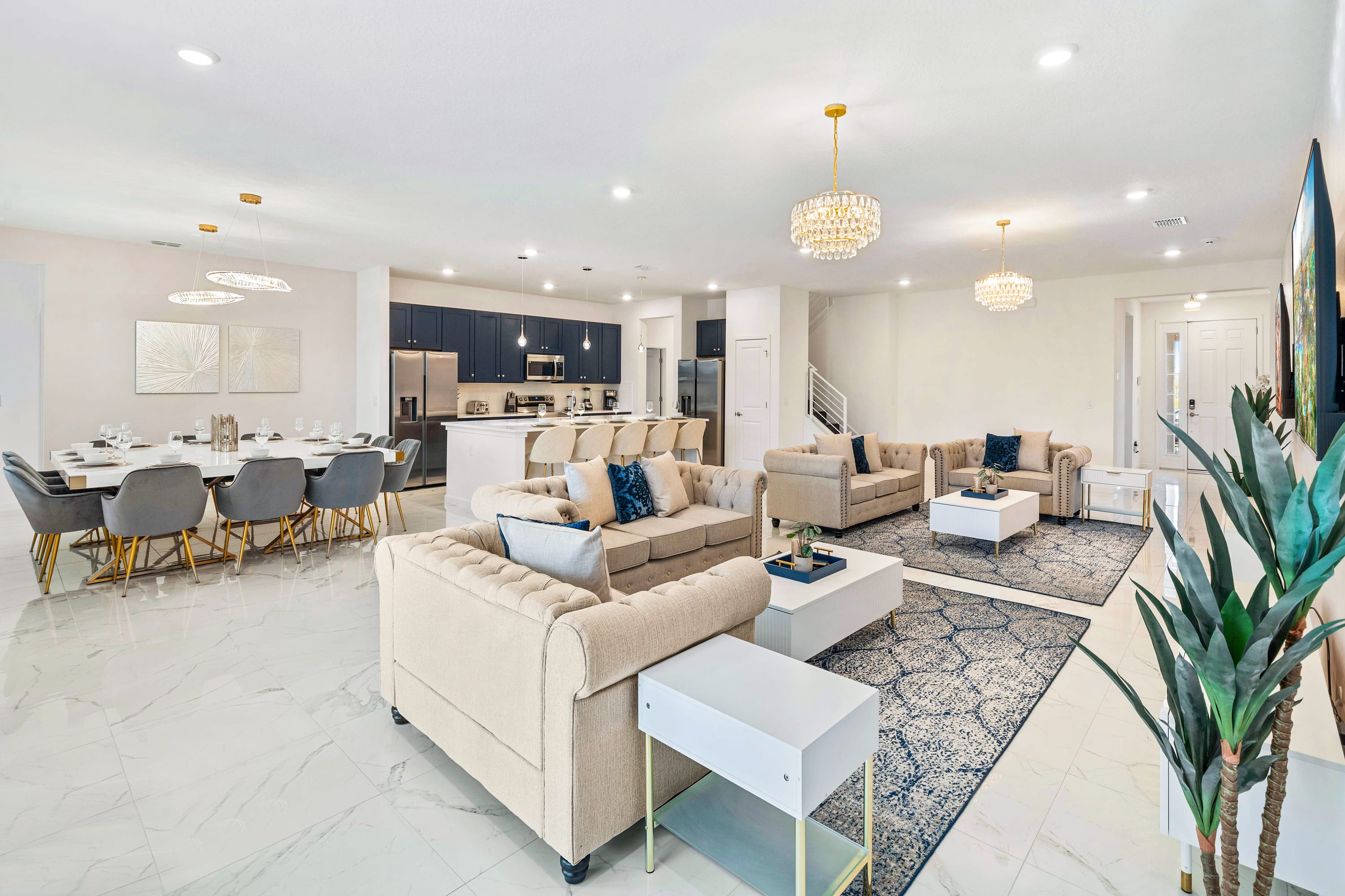 An expansive open-concept living space featuring light beige tufted sofas with blue accent pillows, white coffee tables, and elegant crystal chandeliers, flowing into a dining area and a modern kitchen with dark blue cabinetry.