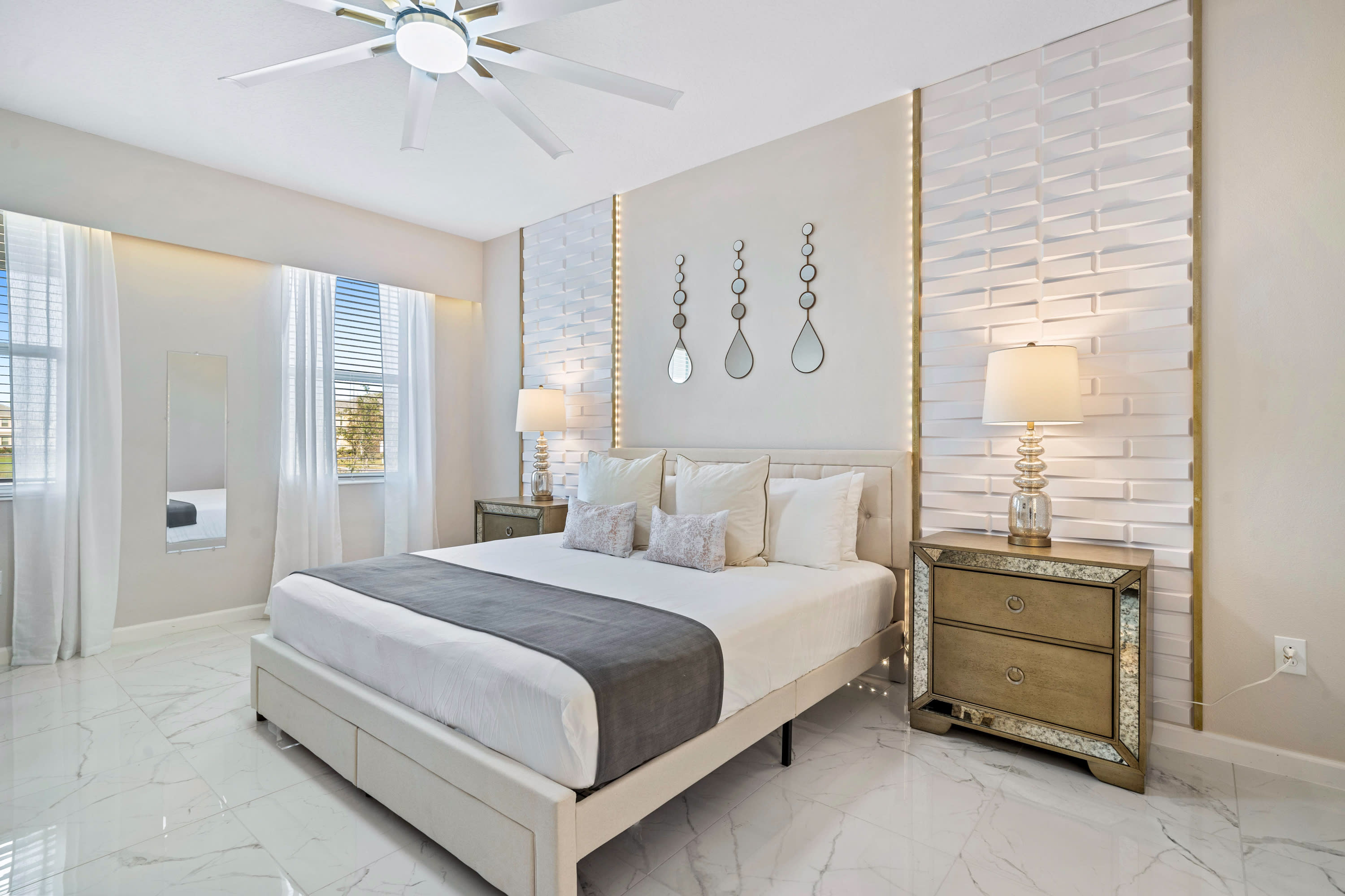 A bright bedroom with white marble-look tile floors, a large bed with a white and grey duvet, and mirrored nightstands. The headboard wall features textured white panels with gold accents and decorative mirrors.