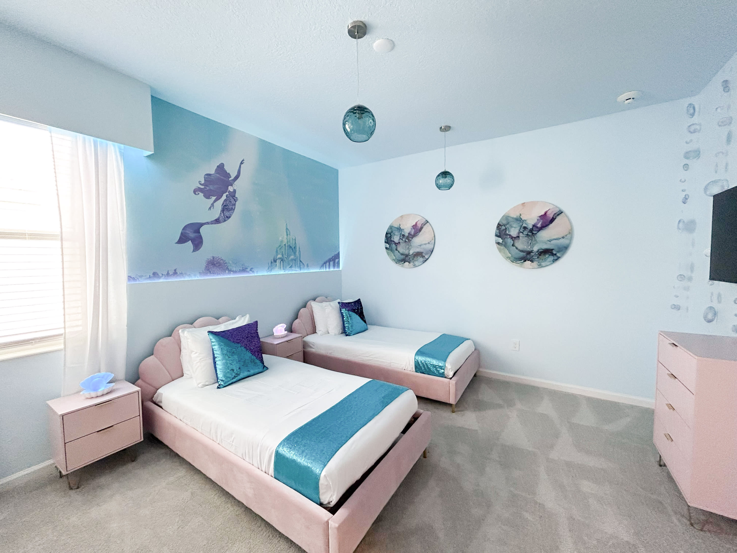 A whimsical children's bedroom featuring two twin beds with pink seashell headboards, white bedding, and blue accents. A large mural of Ariel from The Little Mermaid adorns the light blue wall, complemented by two round abstract art pieces and glass pendant lights.
