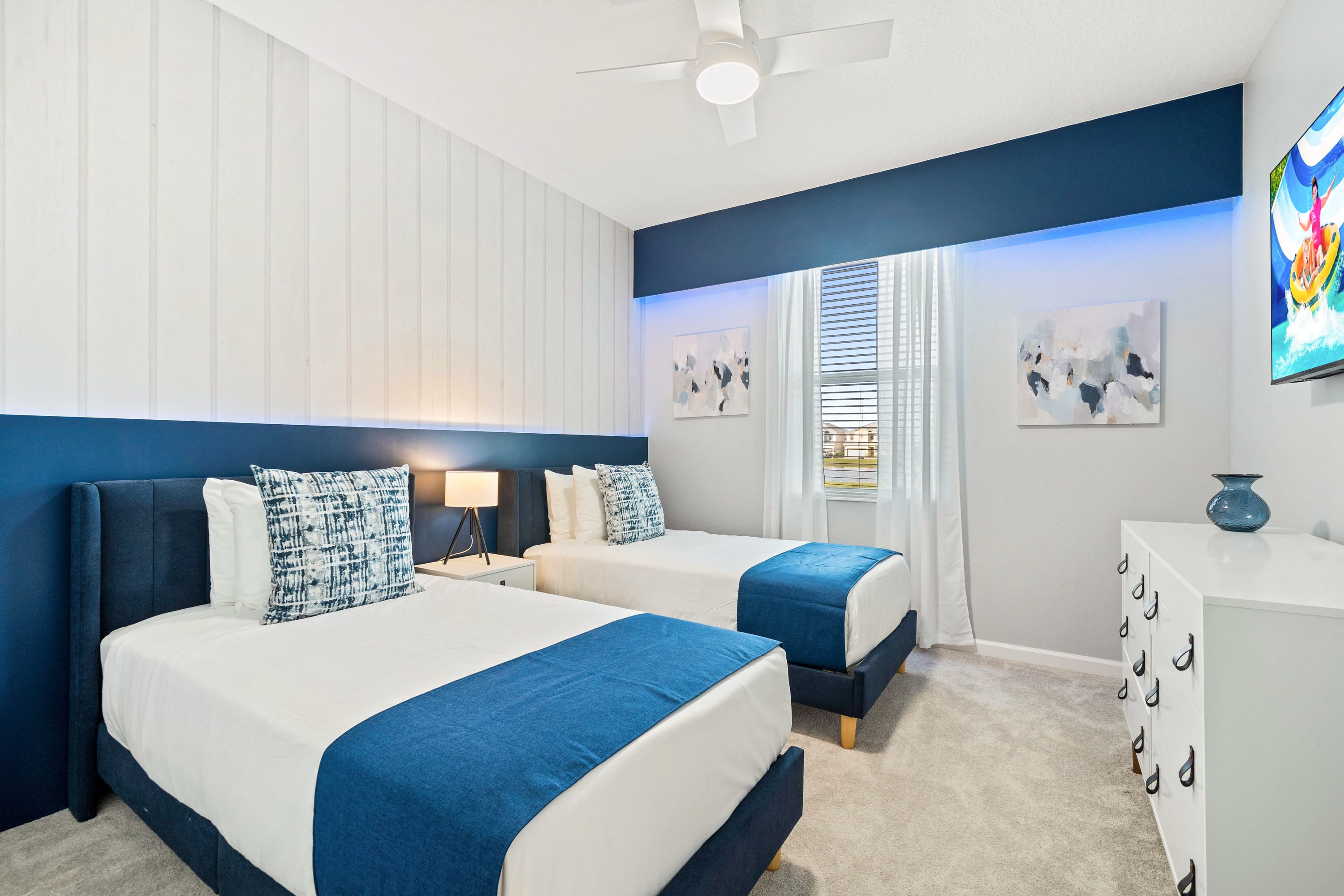 A contemporary bedroom featuring two twin beds with blue upholstered headboards and white bedding accented with blue throws and pillows. The room has white paneled walls, a blue accent wall with integrated lighting, and abstract artwork, creating a serene ambiance.