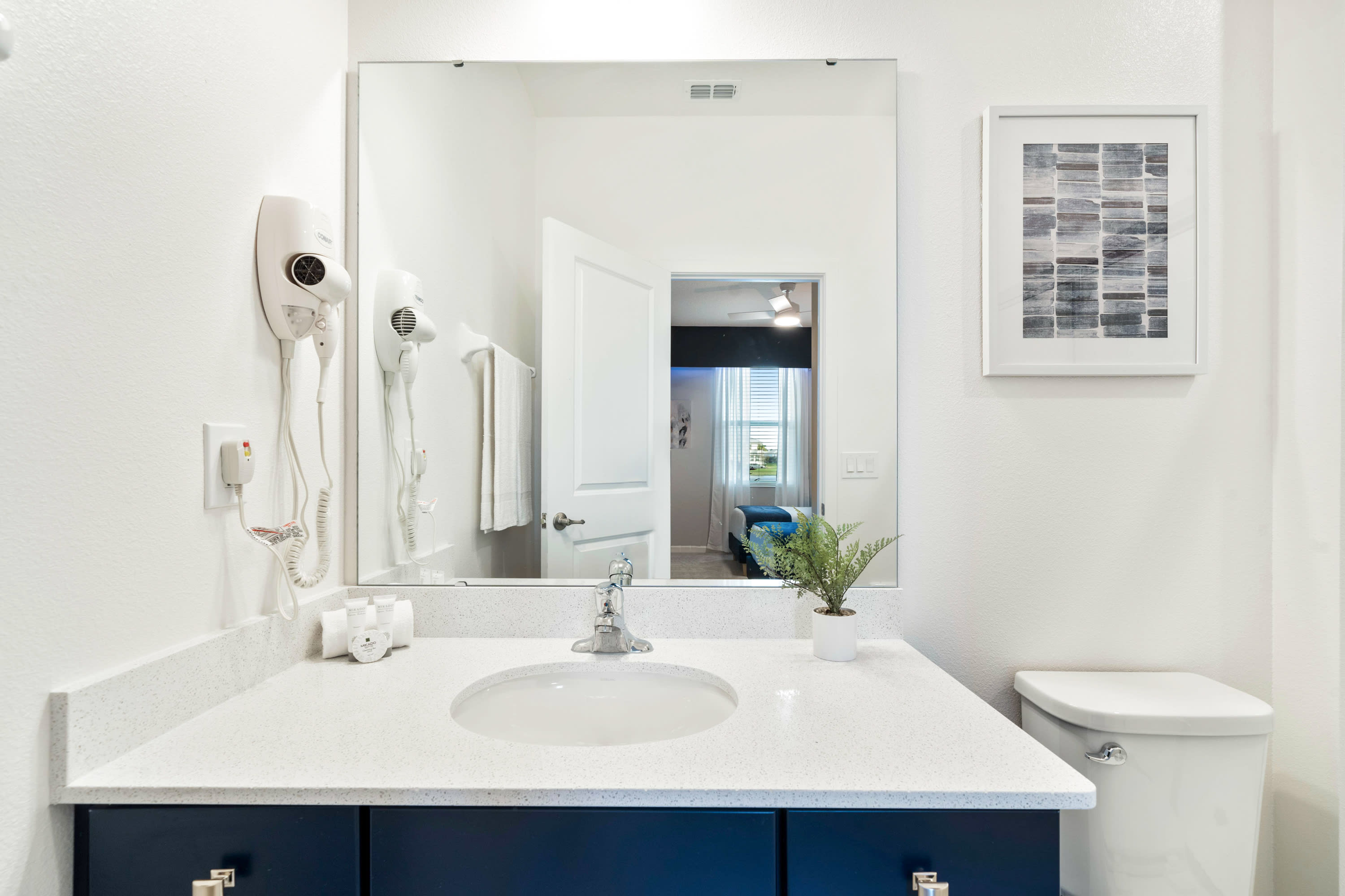 Clean and functional bathroom vanity area with a single sink set in a white countertop with a dark blue cabinet. Two hair dryers are mounted on the wall next to the large mirror, which reflects the adjacent bedroom.