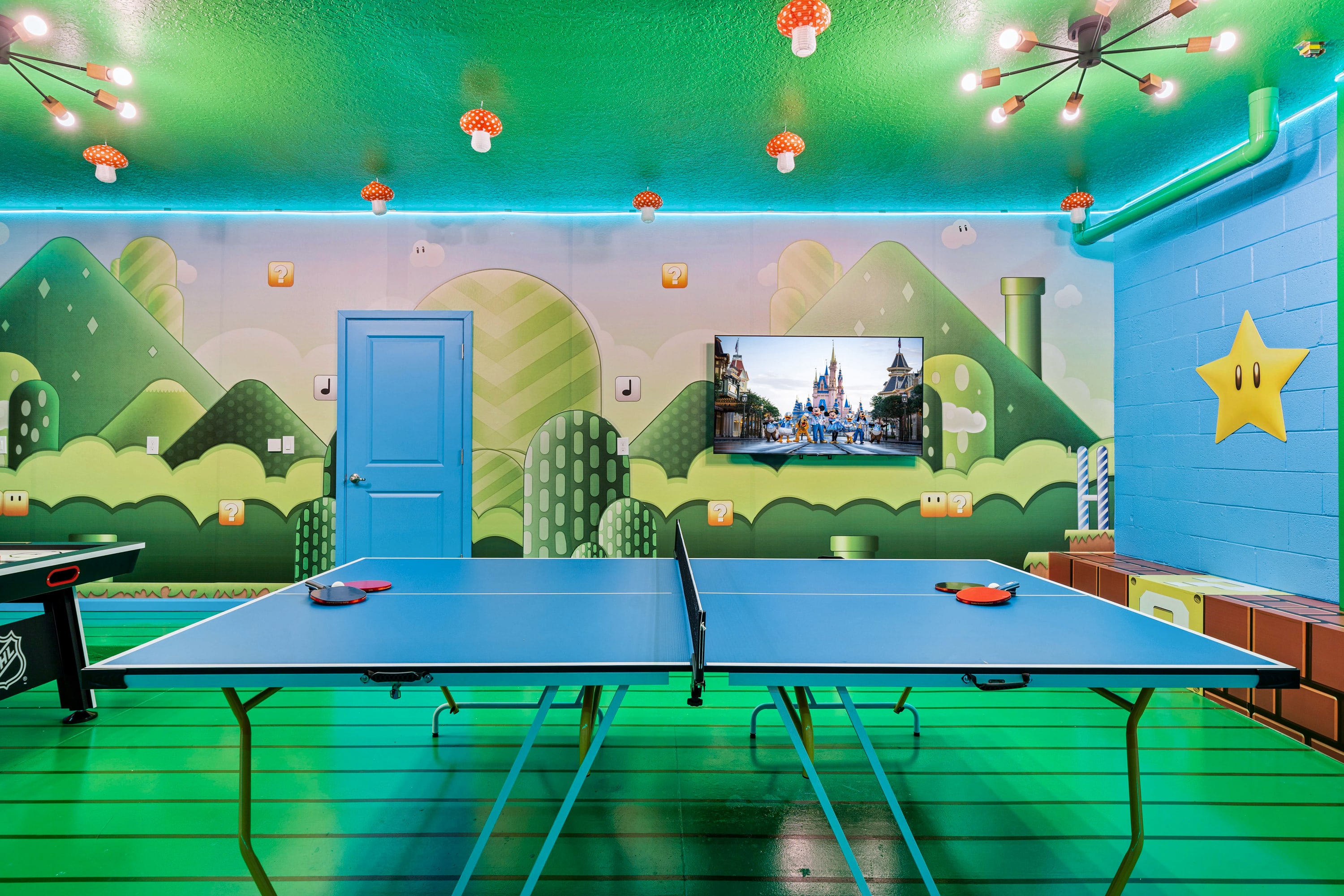 Brightly lit game room with a Super Mario Bros. theme, featuring a blue ping pong table, a wall-mounted TV, and green-patterned walls and floor, with mushroom-shaped ceiling lights.