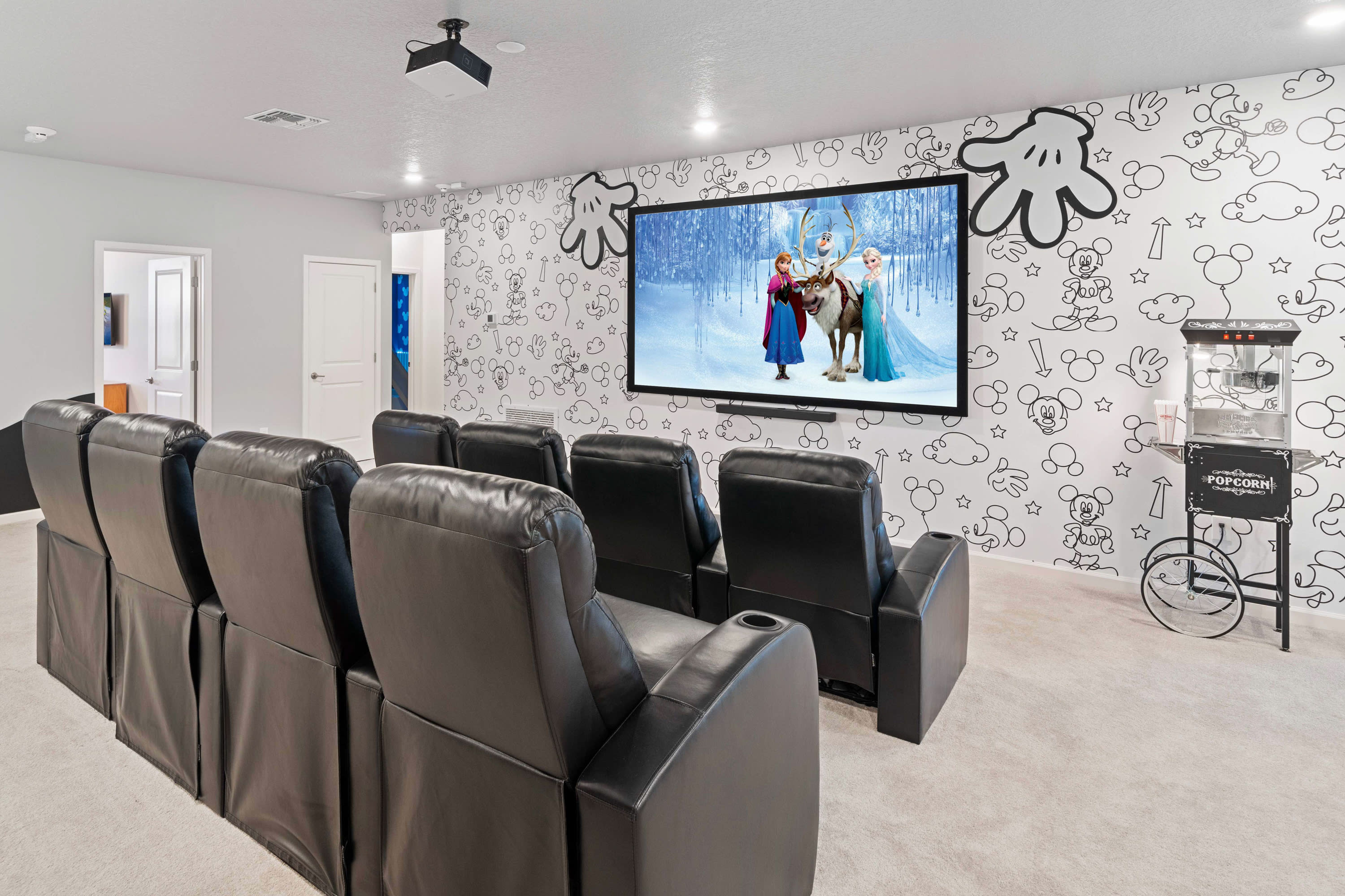 A home theater with a large projection screen displaying an animated movie, featuring multiple rows of black leather reclining seats and a popcorn machine, all set against a whimsical cartoon-themed wall.