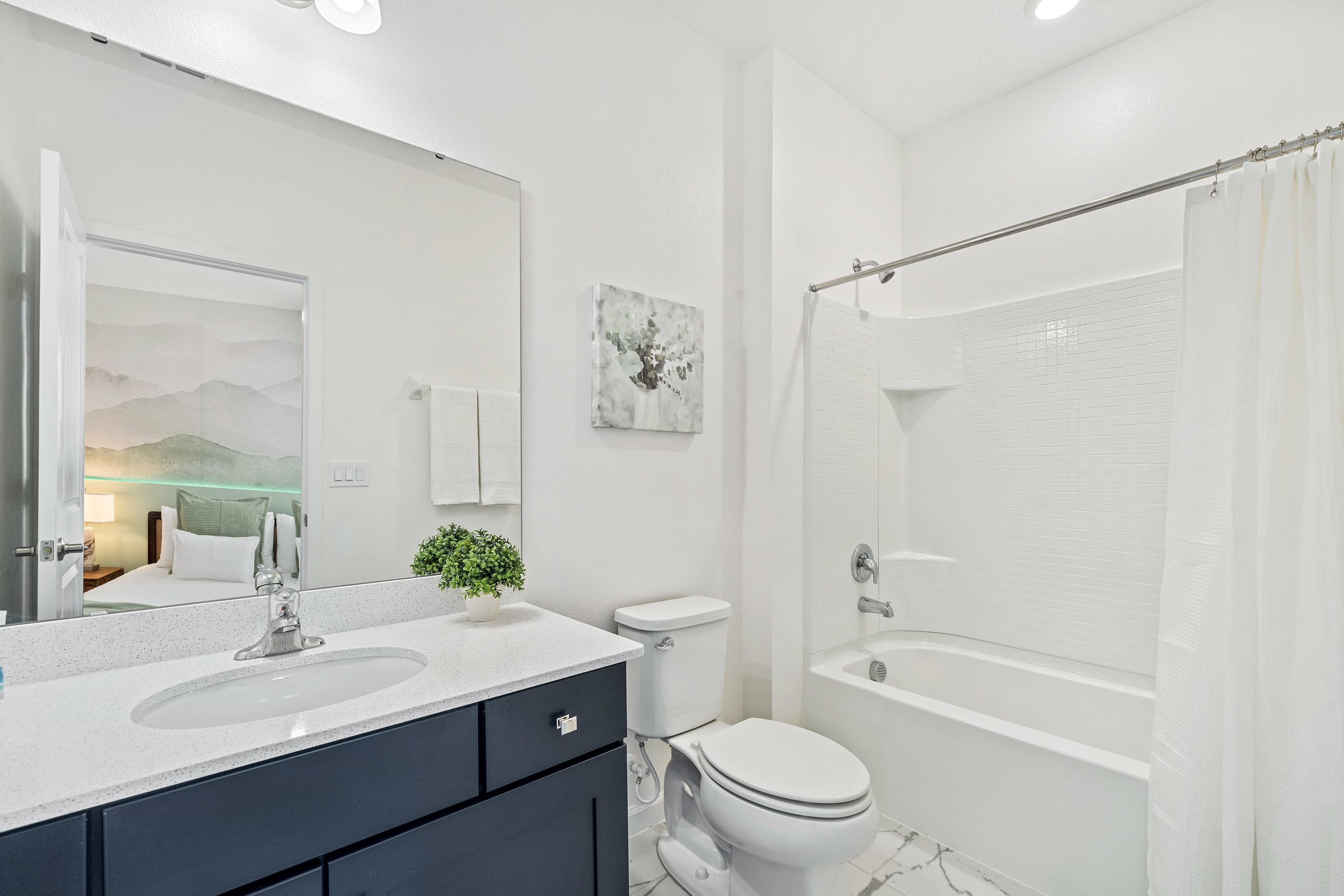 Clean white bathroom with a dark blue vanity, white countertop, and single sink, alongside a toilet and a shower-tub combo with white subway tile. A small green plant adorns the vanity, and a mirror reflects a bedroom with a green mountain mural.