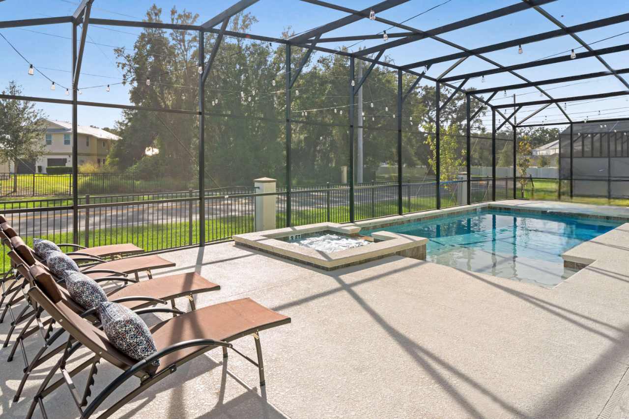 Screened outdoor pool area with a rectangular swimming pool and an adjacent hot tub, surrounded by several lounge chairs on a light-colored concrete deck, overlooking a fenced backyard.
