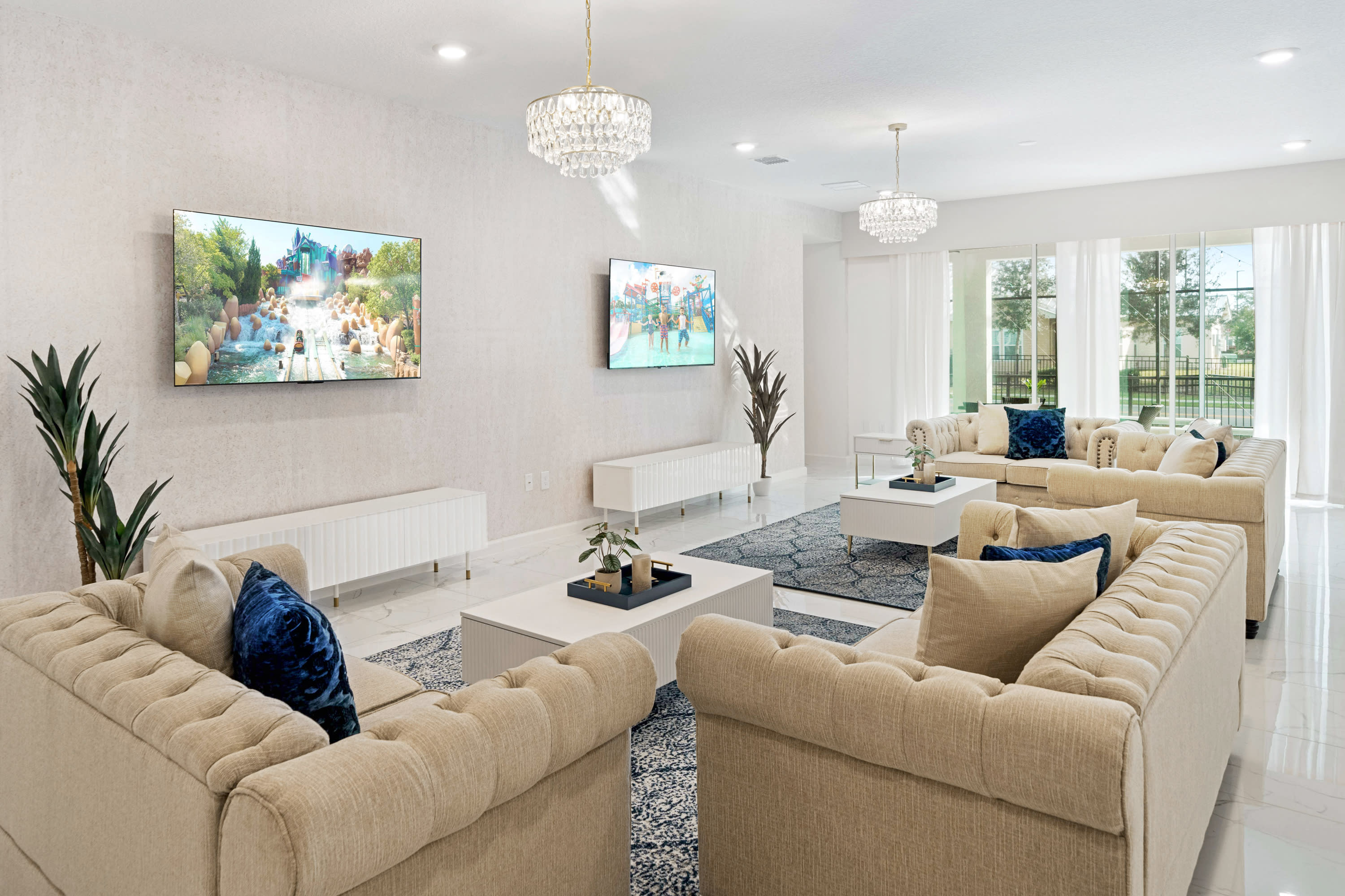 A bright and spacious living room featuring two large beige tufted sofas with blue velvet pillows, two white coffee tables, and dual wall-mounted TVs, illuminated by crystal chandeliers and natural light from large windows.