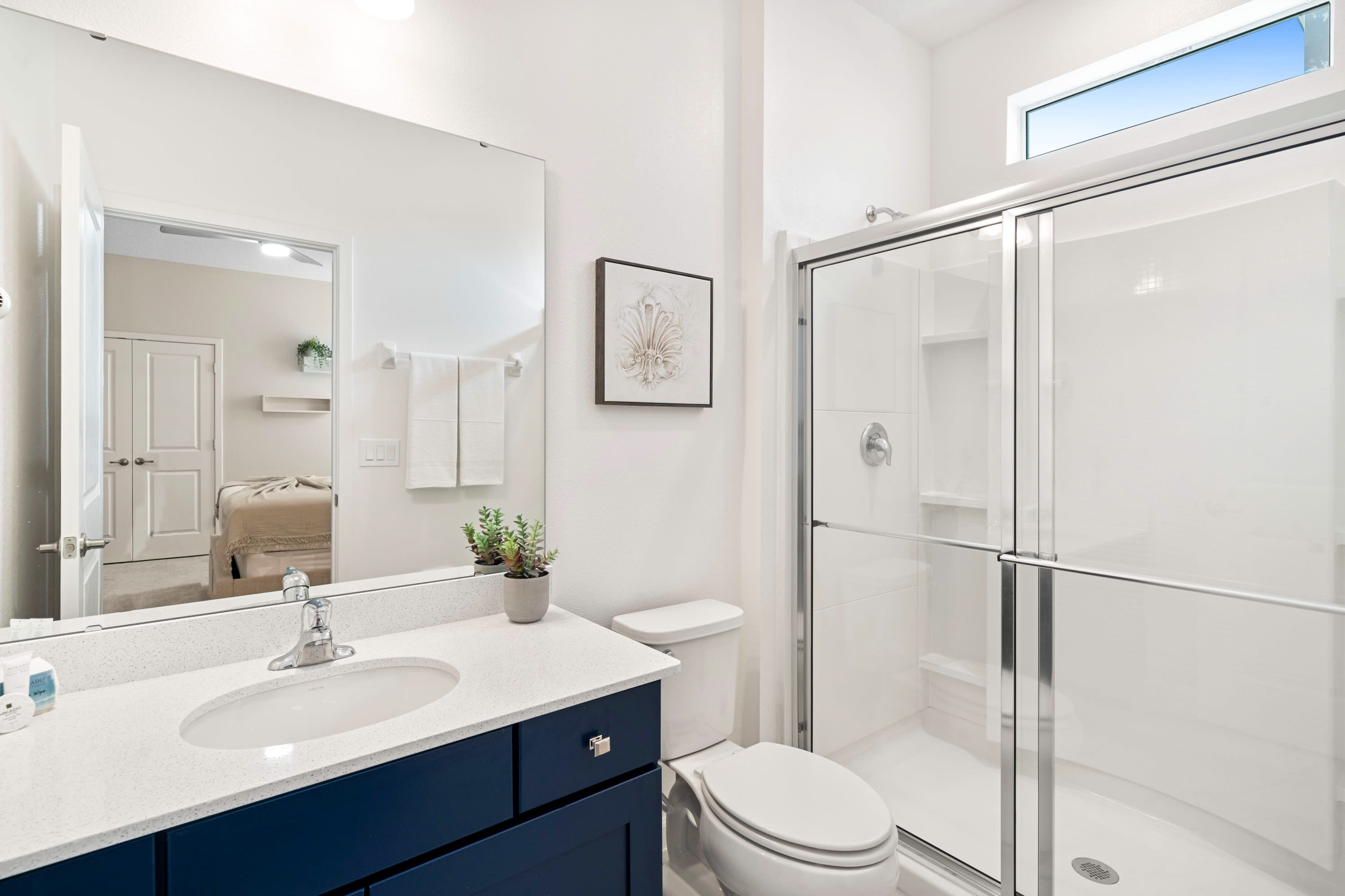 A modern white bathroom with a single dark blue vanity, white countertop, and a large mirror. It includes a toilet and a glass-enclosed walk-in shower, with a high window for natural light.