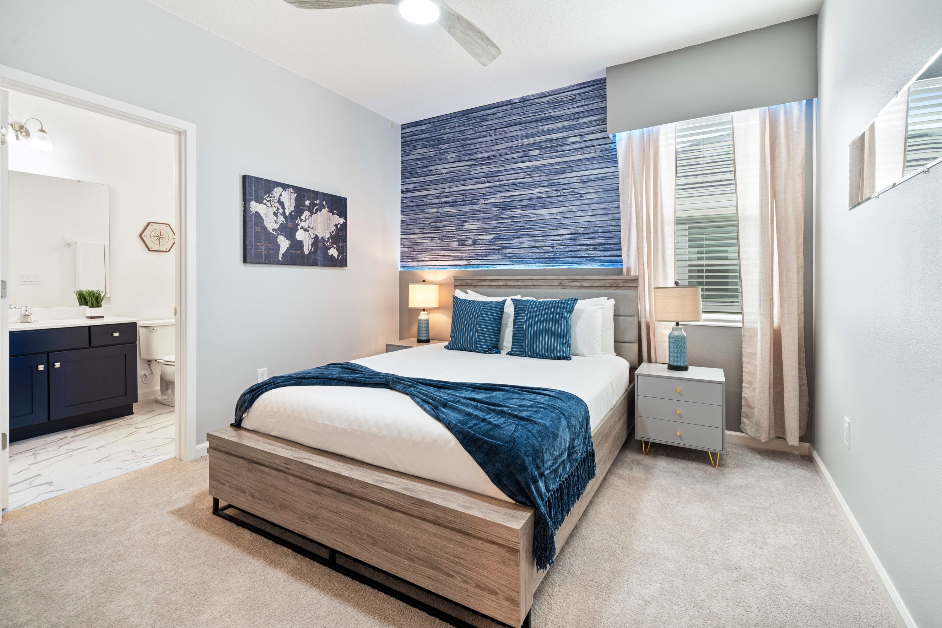 Modern bedroom featuring a king-sized bed with a light wood frame and blue bedding, complemented by a textured blue accent wall behind the headboard. Two grey nightstands with blue lamps flank the bed, and a ceiling fan is overhead.