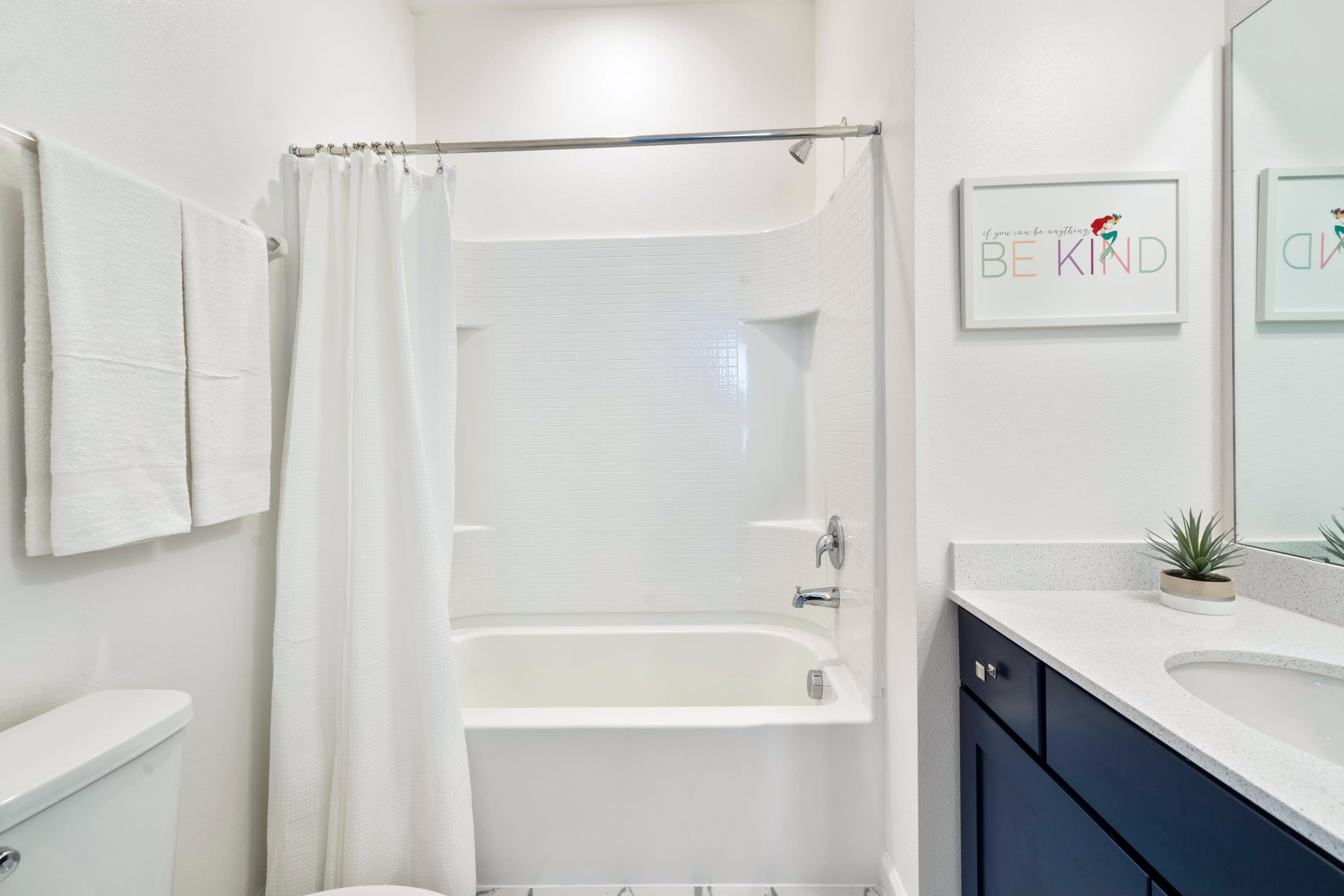 Bright white bathroom with a tiled tub/shower combo and a single vanity featuring a dark blue cabinet and white countertop. A framed 'BE KIND' art piece with a mermaid tail accent hangs above the toilet, adding a playful touch.