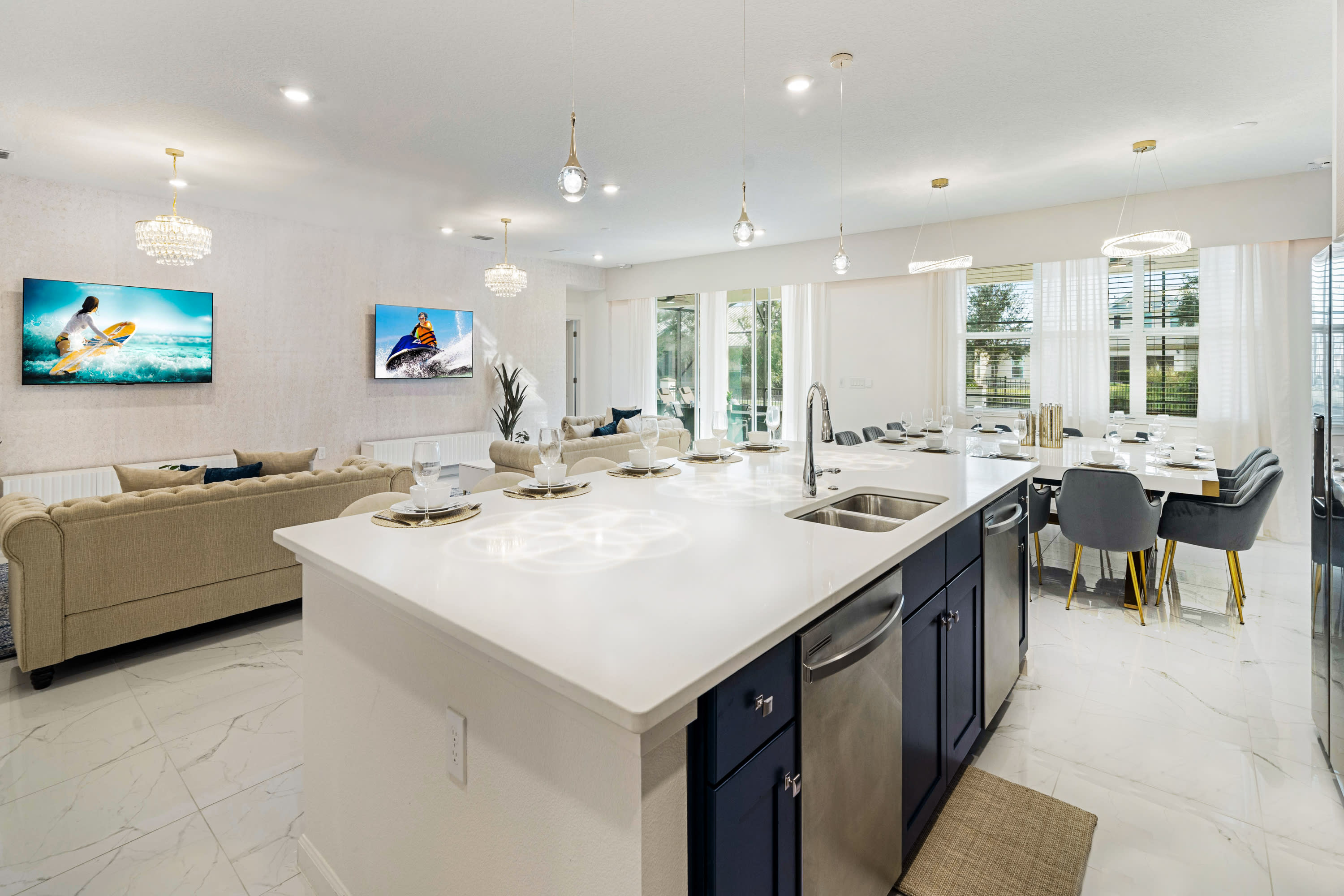 A modern kitchen featuring a large white island with a sink and white bar stools, dark blue lower cabinetry, and stainless steel appliances, with the dining and living areas visible in the background.