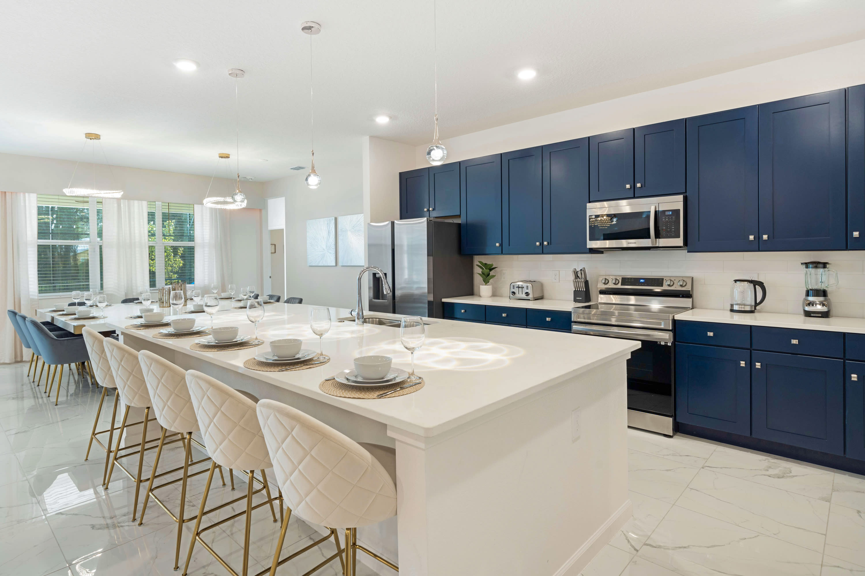 A sleek kitchen showcasing dark blue cabinetry with white quartz countertops and a white subway tile backsplash, equipped with stainless steel appliances and a large island featuring white bar stools and pendant lighting.