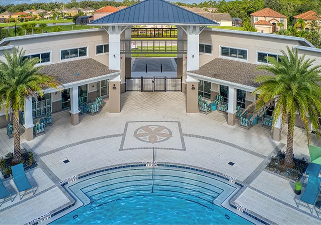 An aerial view of a large communal pool area featuring a multi-level pool with steps and a patterned tile deck. A modern clubhouse building with a distinctive roof and palm trees surrounds the inviting space.