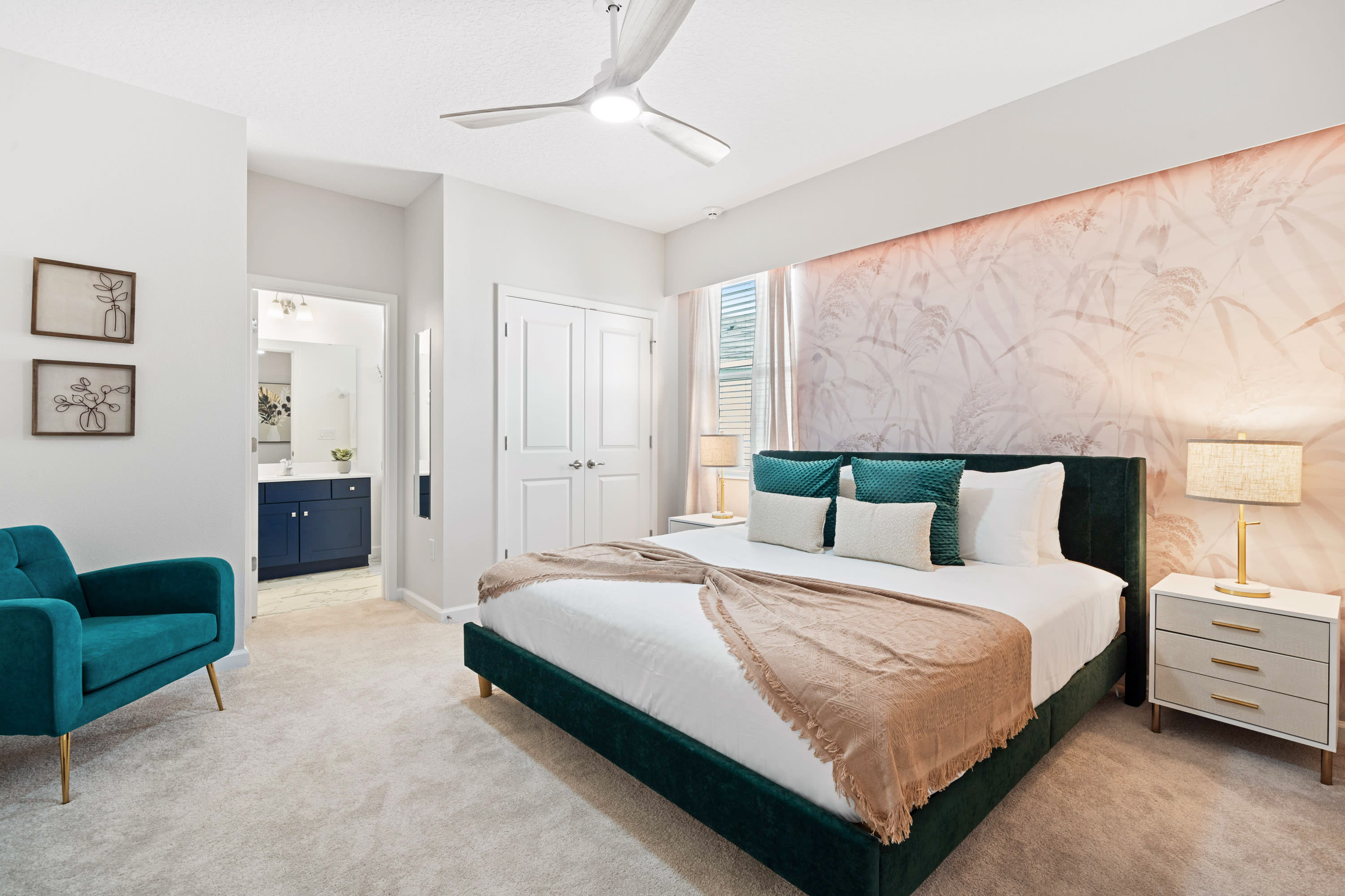 Elegant bedroom featuring a king-sized bed with a green velvet headboard and white bedding, set against a soft pink botanical wallpaper accent wall. A teal armchair and white nightstands with lamps complete the inviting space.
