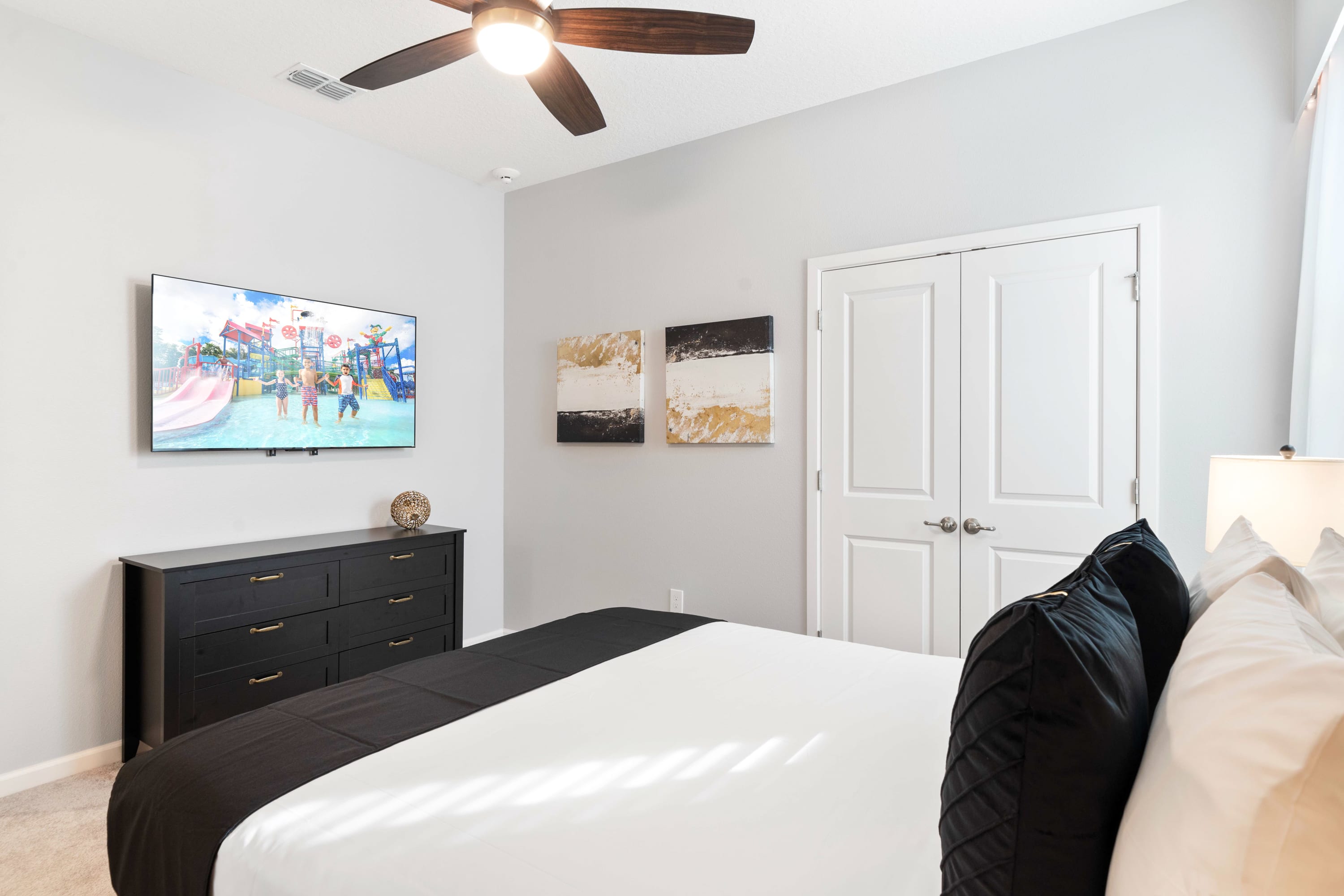 Cozy bedroom with a queen-sized bed featuring black and white bedding, a black dresser with a mounted TV, and abstract black and gold artwork on the wall. A ceiling fan and white closet doors are also visible.