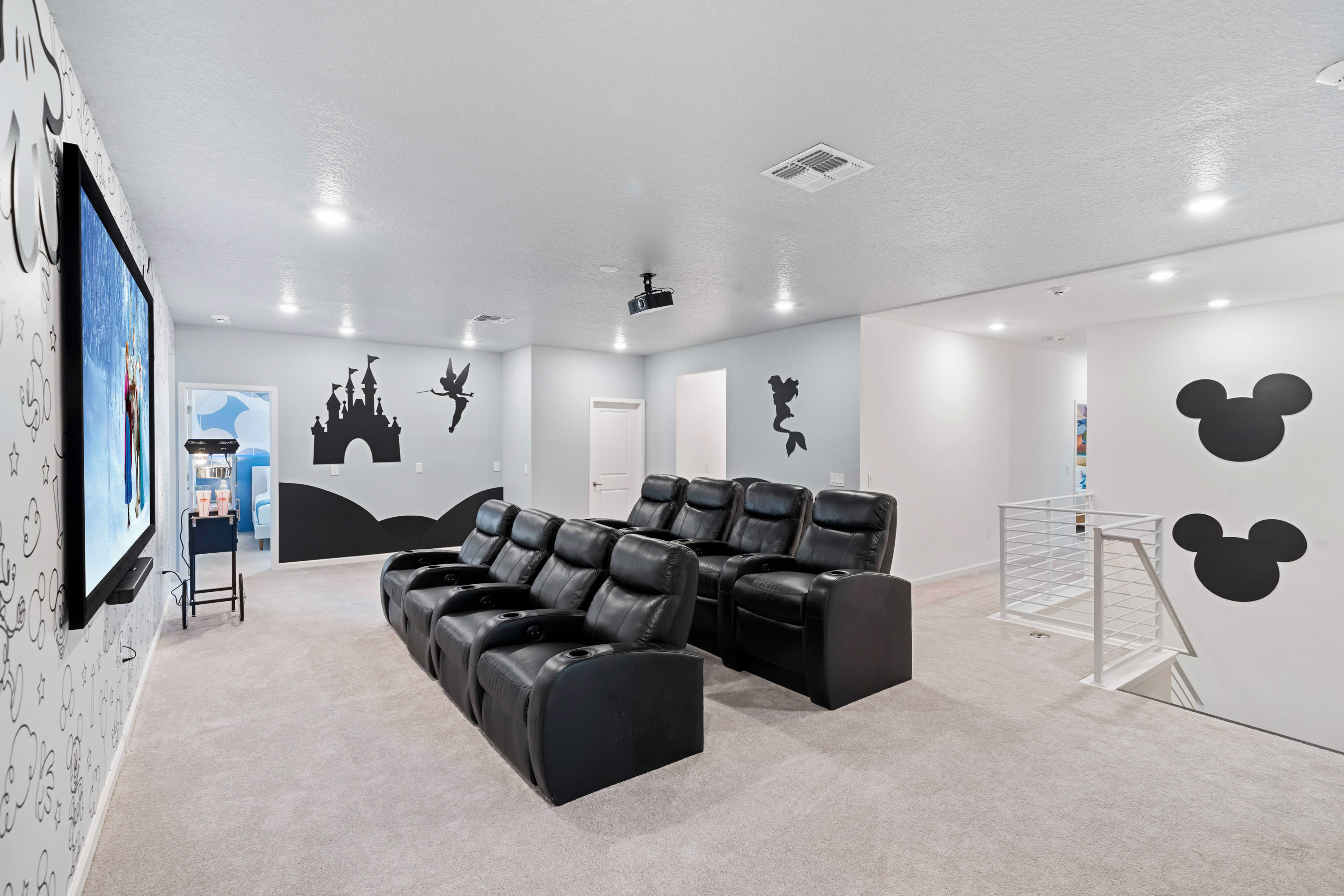 A spacious home theater room with two rows of black leather reclining seats facing a large screen, featuring a ceiling-mounted projector and walls adorned with whimsical castle, fairy, and mermaid silhouettes.