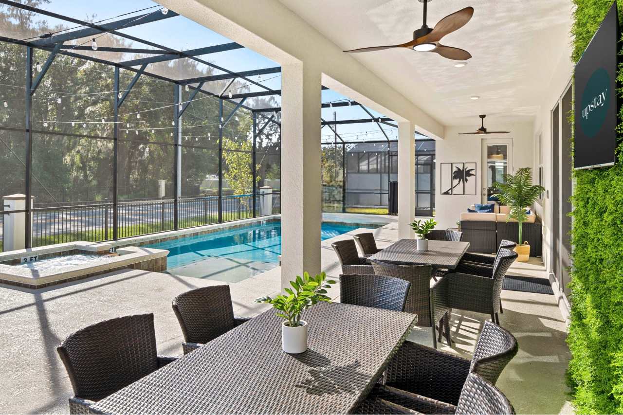 Covered outdoor patio within a screened enclosure, featuring multiple wicker dining tables and chairs, ceiling fans, and a view of the swimming pool and hot tub.