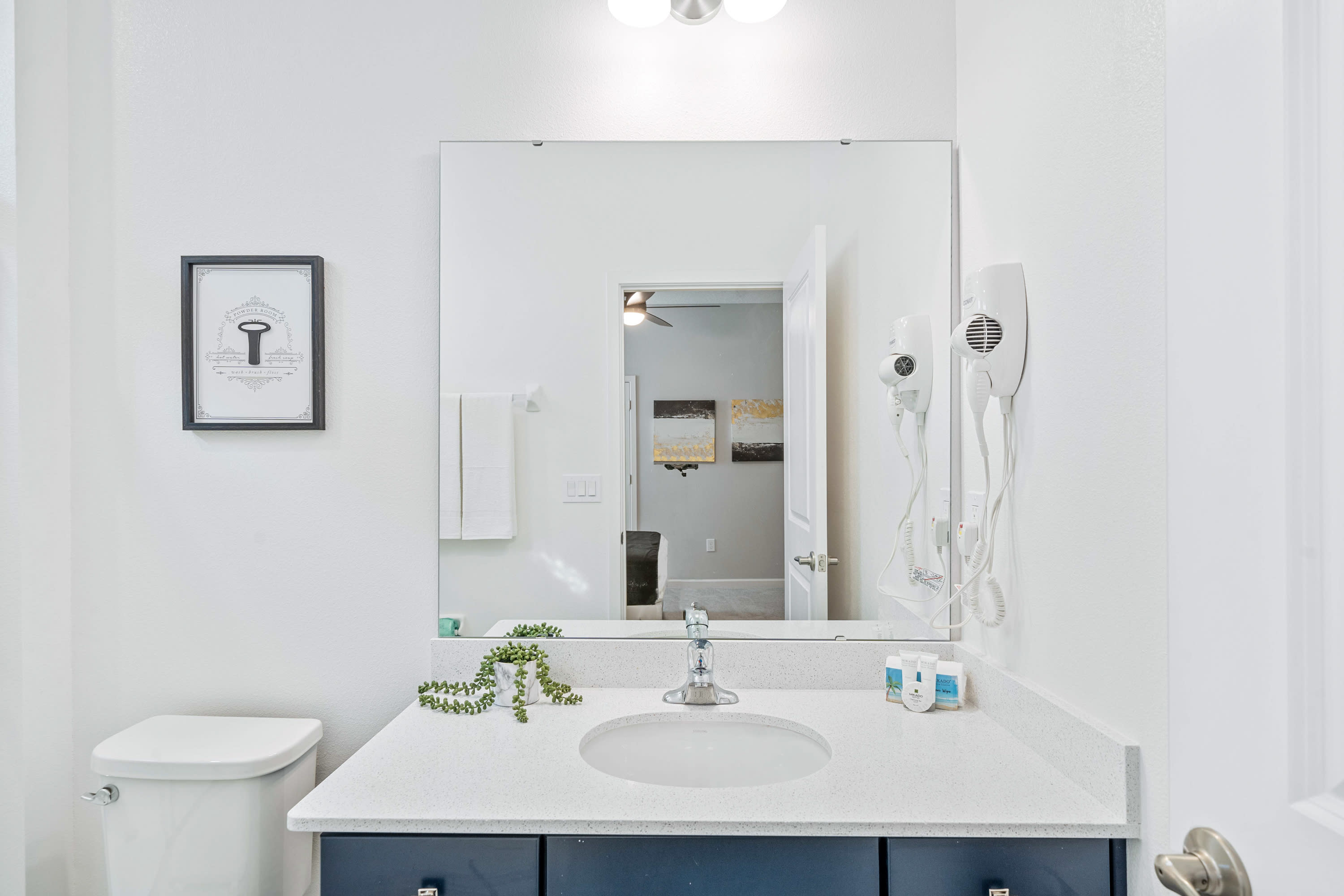 Clean white bathroom with a dark blue vanity, white countertop, and single sink, a toilet, and a shower-tub combo. A framed black and white print hangs on the wall, and a mirror reflects a bedroom with abstract art.