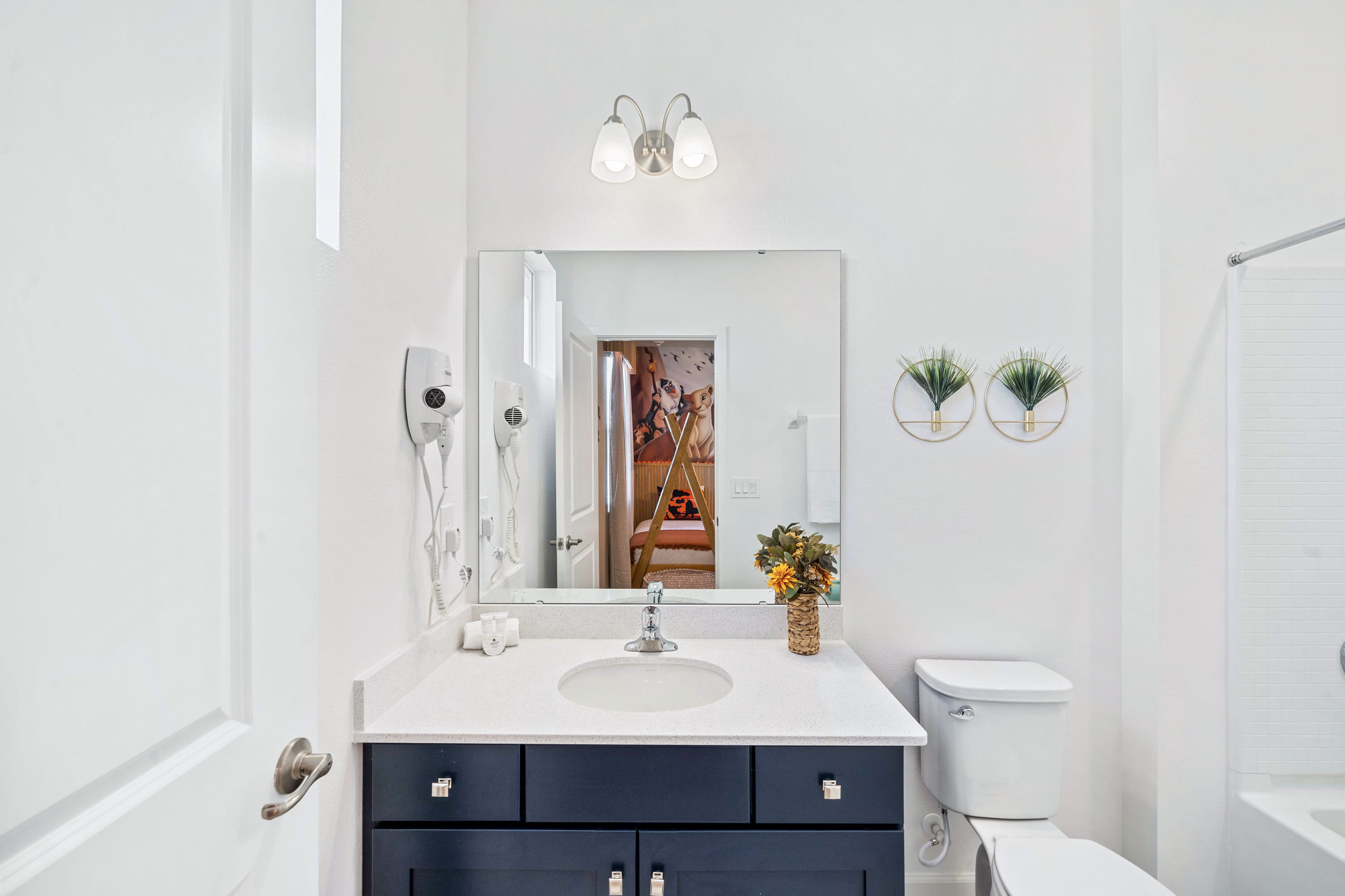 Bright white bathroom with a dark blue vanity, white countertop, and single sink, a toilet, and a shower-tub combo. Two gold geometric wall decorations with plants hang above the toilet, and a mirror reflects a Lion King themed bedroom.